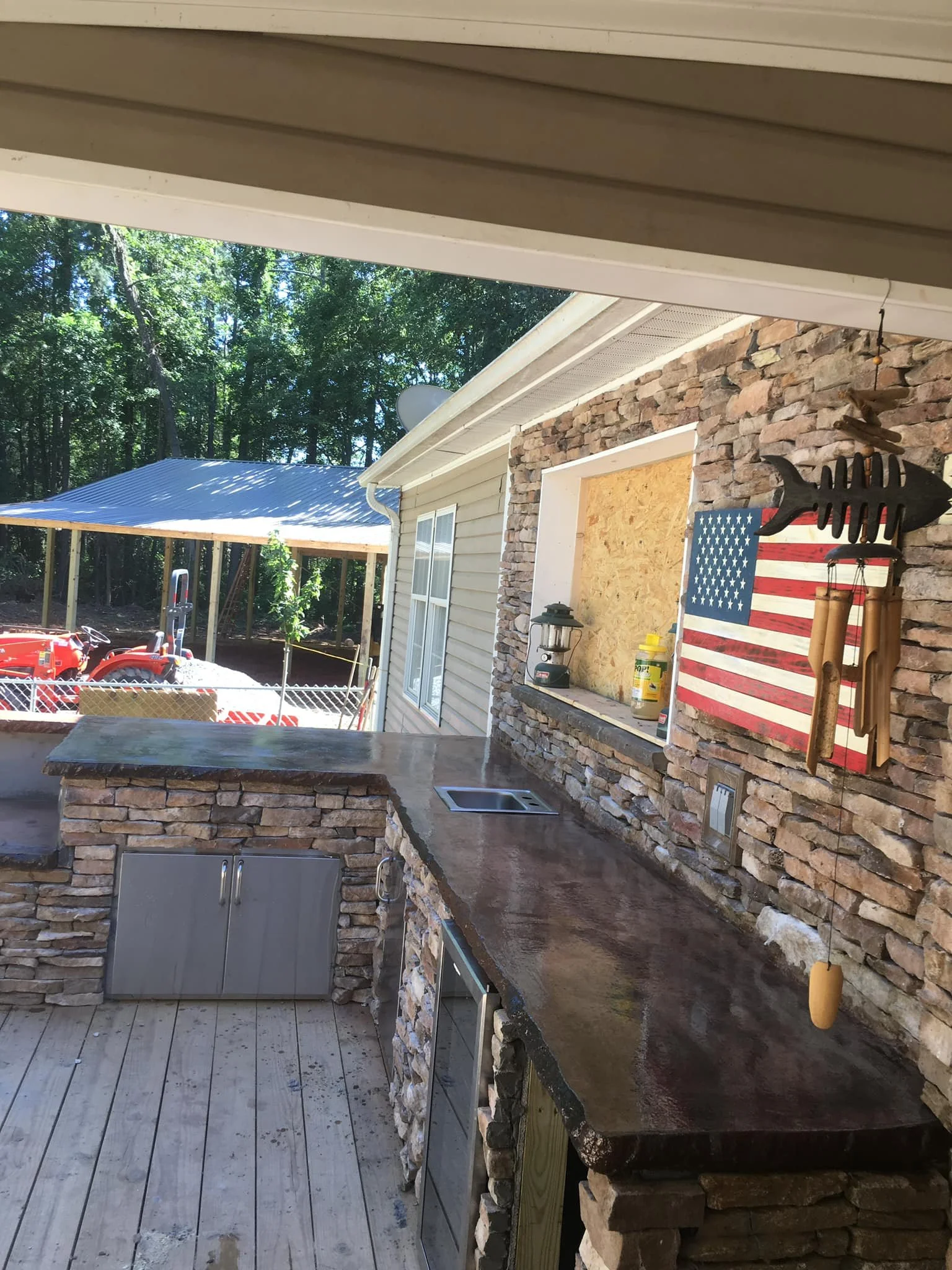 Outdoor area with a stone countertop and backsplash, an American flag wall decor, and a small window with a board covering it. A lantern, spray bottle, and wooden fish decoration hang on the wall. In the background, there is a red tractor, a shed wit