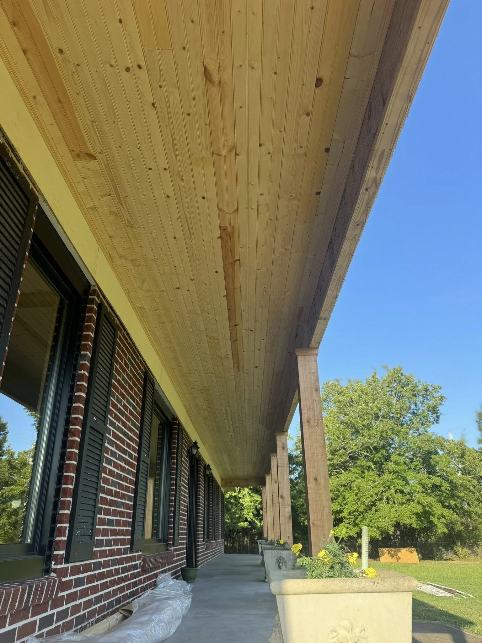 The image shows a newly constructed brick building with black shutters, a concrete pathway, and a large wooden porch with supporting columns. There are planters with yellow flowers along the pathway, and trees and a clear blue sky are visible in the 