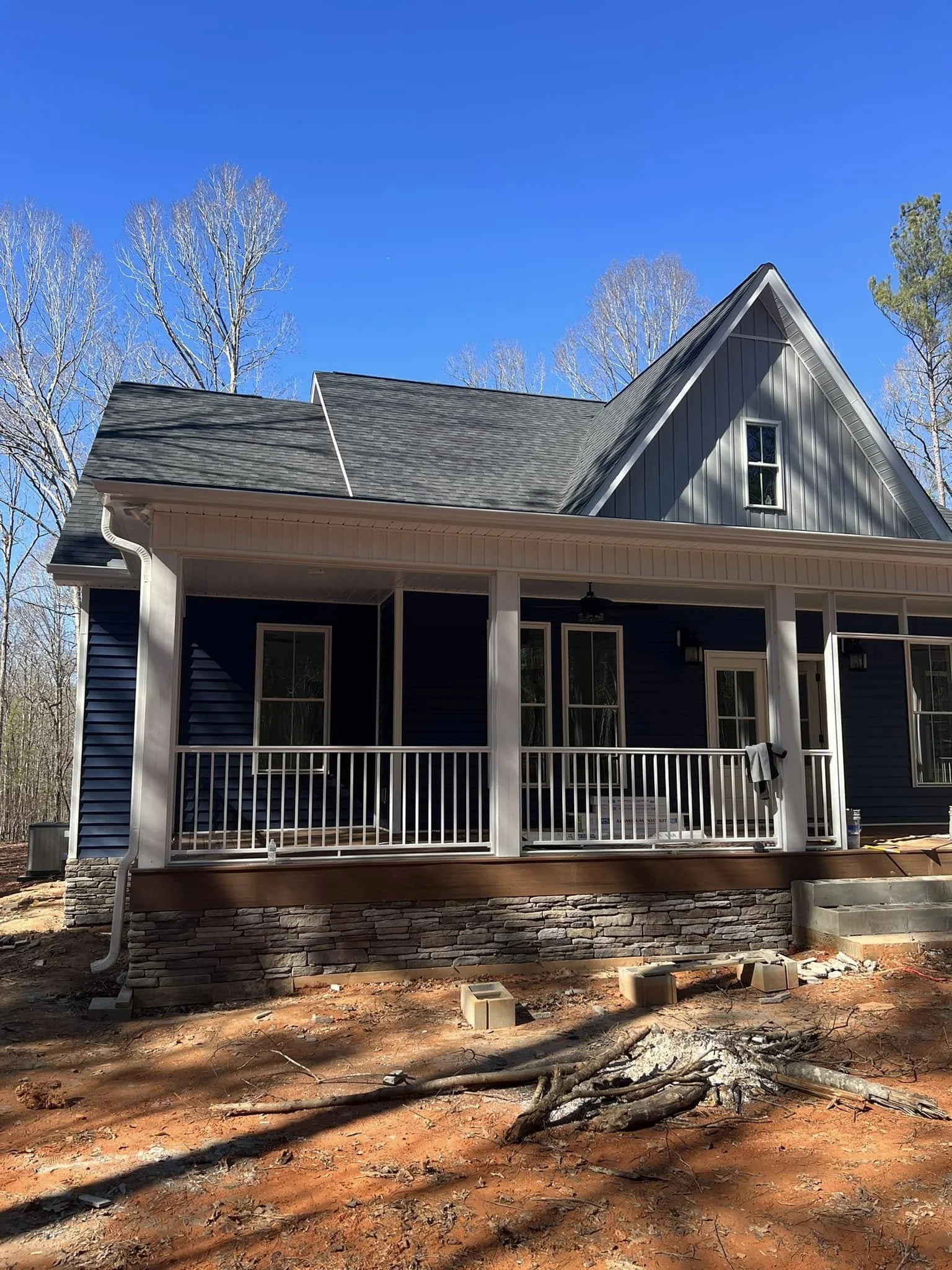 Newly constructed blue house with a deck, stone foundation, and a roof with gables, surrounded by leafless trees under a clear blue sky.