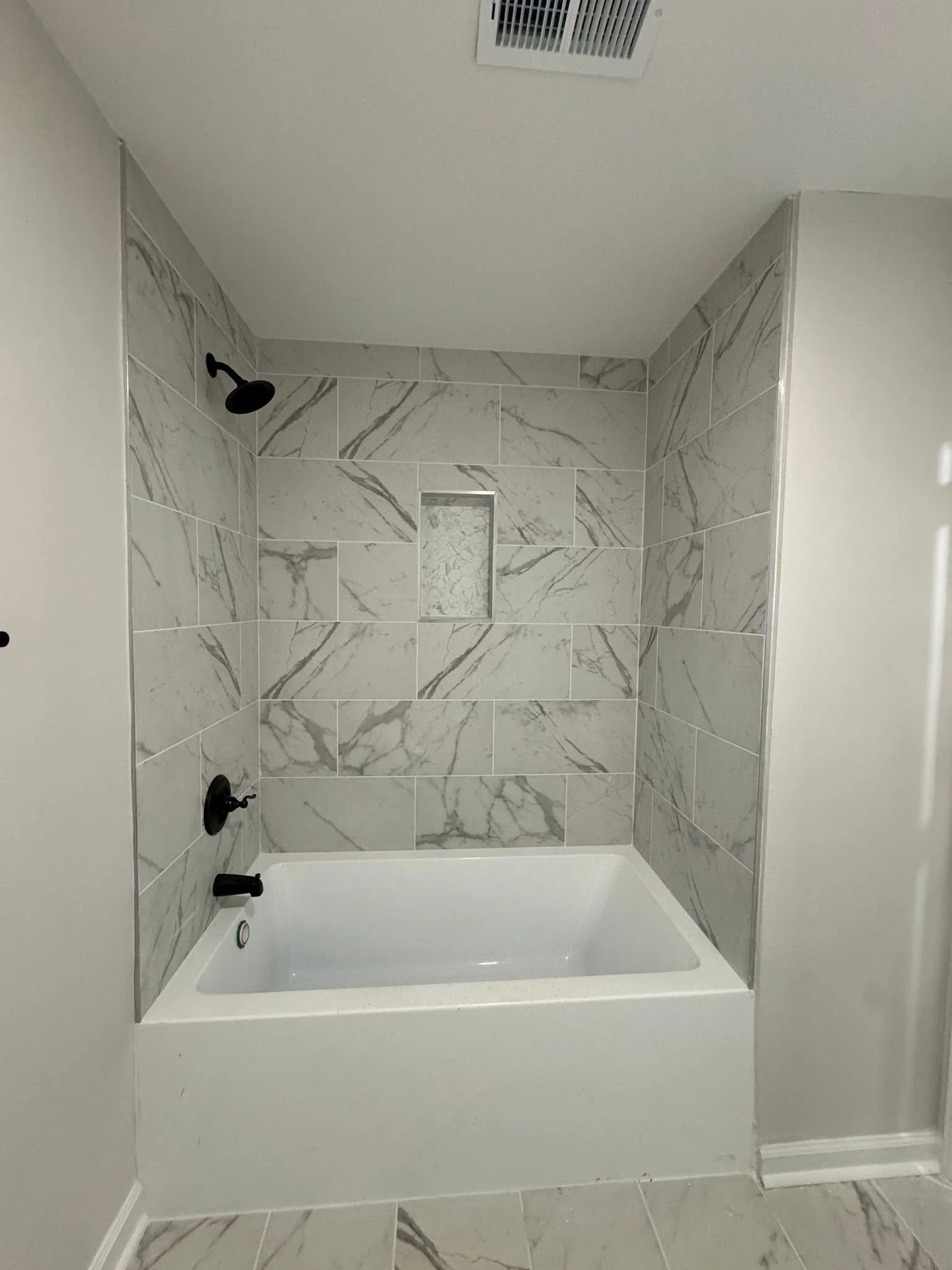 A modern bathroom shower with gray marble tiles, a built-in niche, black shower fixtures, and a white bathtub.