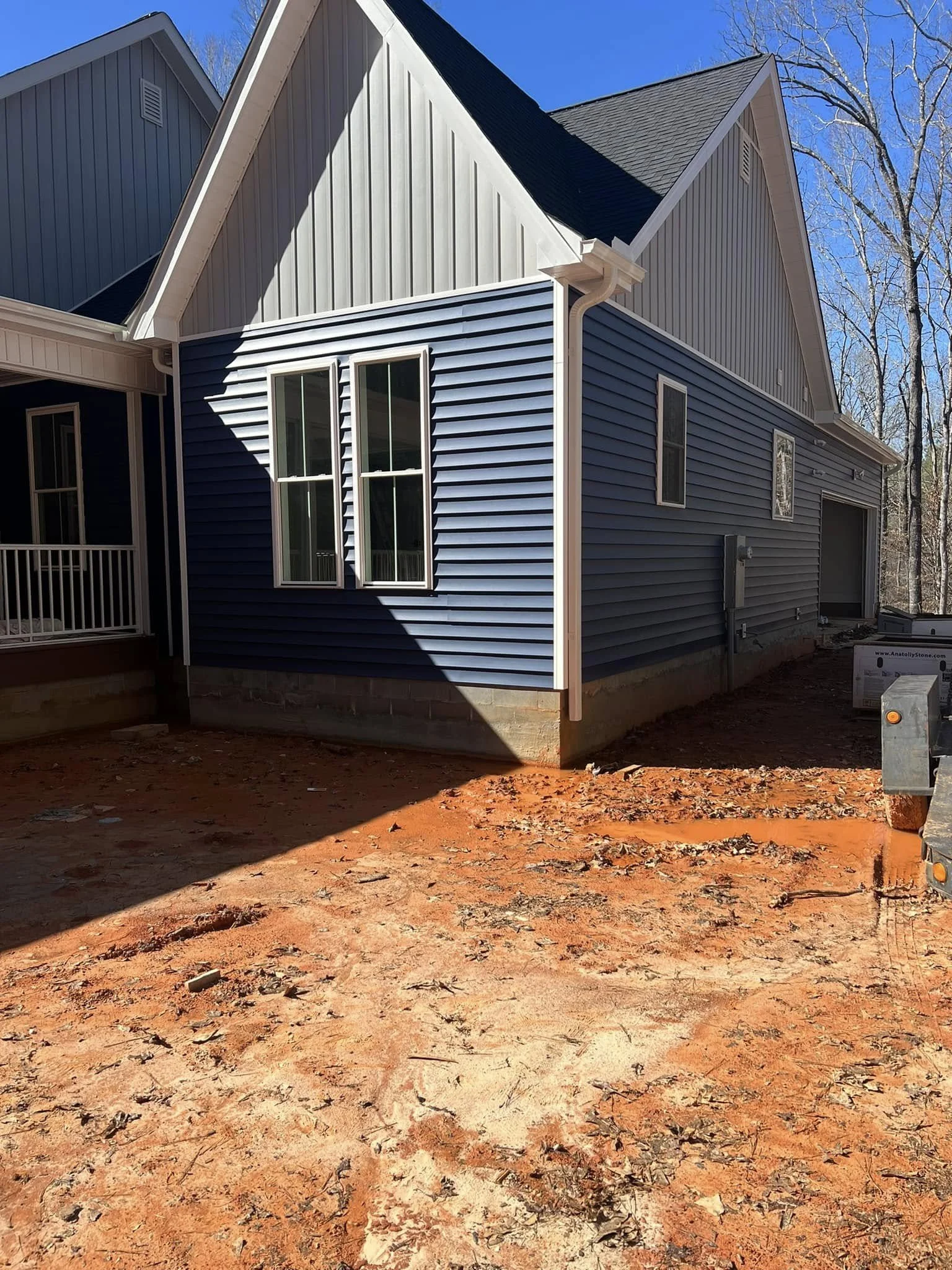Newly constructed house with blue siding, white trim, and a black roof, with a dirt area in the front yard and trees in the background.