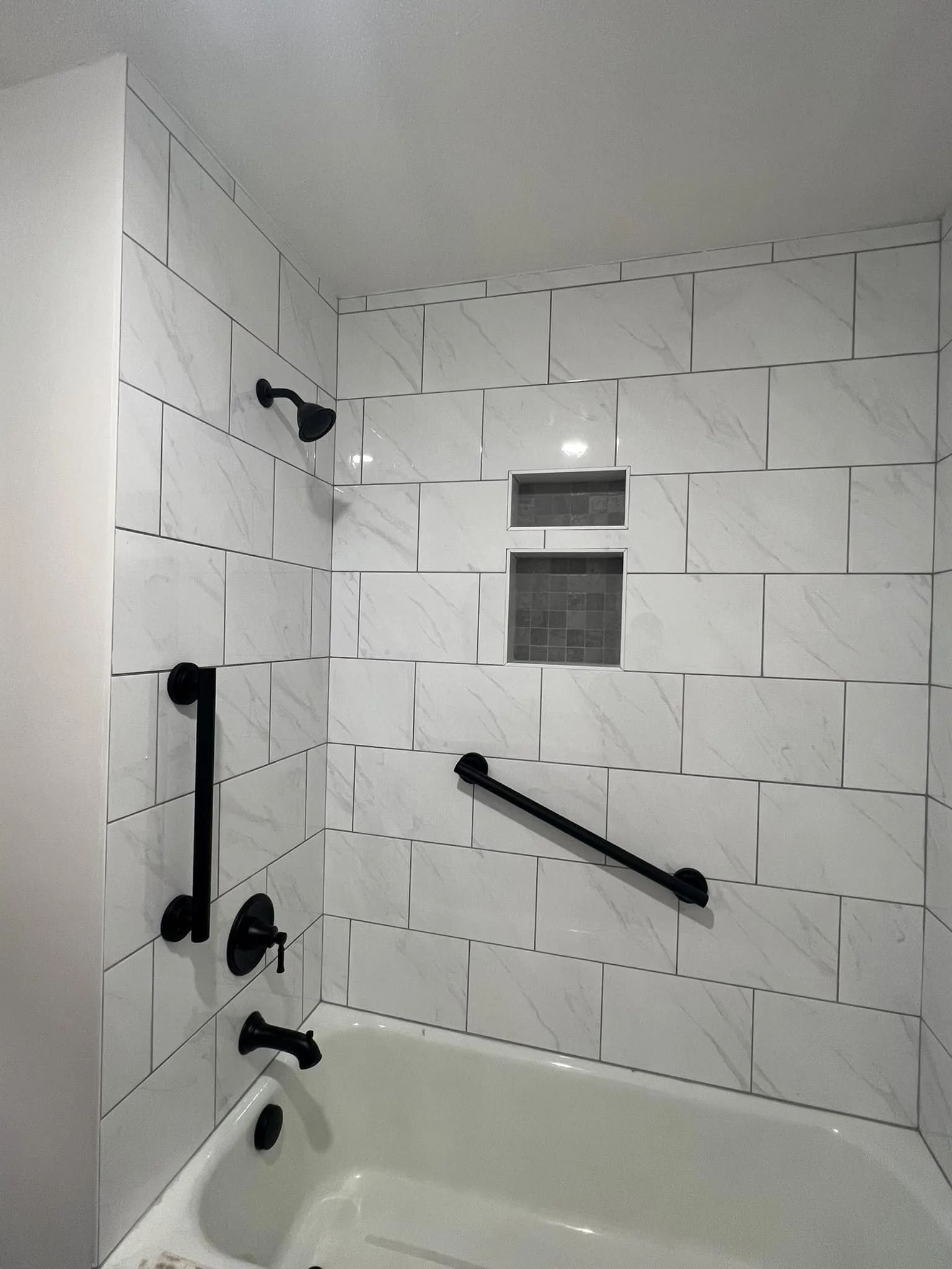 Bathroom shower with white marble tiles, black fixtures, two built-in niche shelves, and a black grab bar.