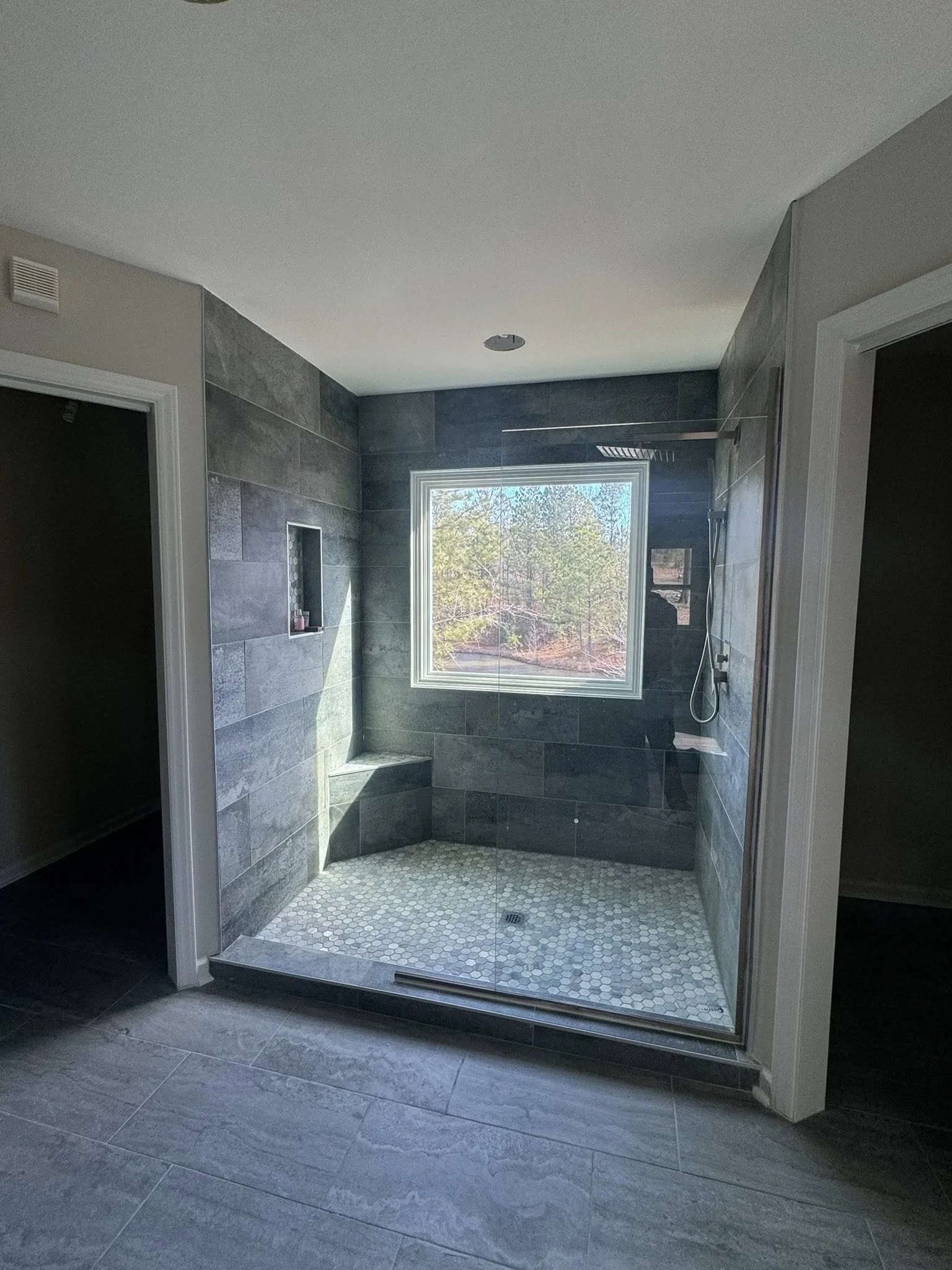 Modern walk-in shower with gray tiles, a built-in niche, a bench, patterned pebble floor, and a large window showing trees outside.