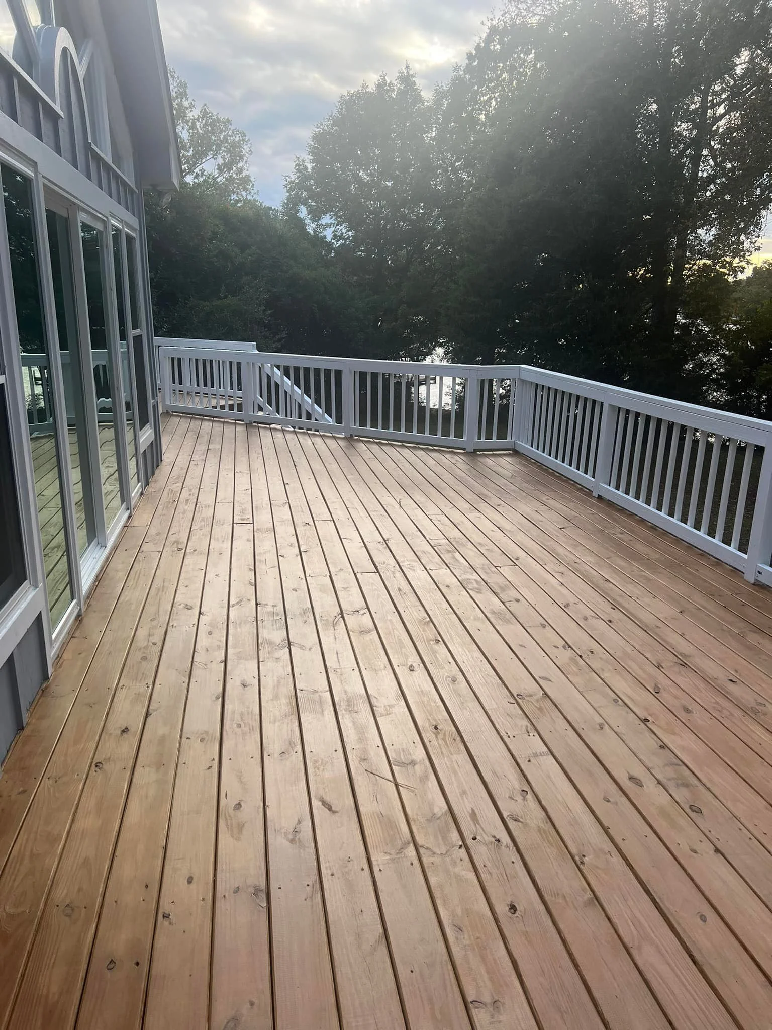 Empty wooden deck with white railing, attached to a house with large glass sliding doors, next to tall trees, and a cloudy sky overhead.
