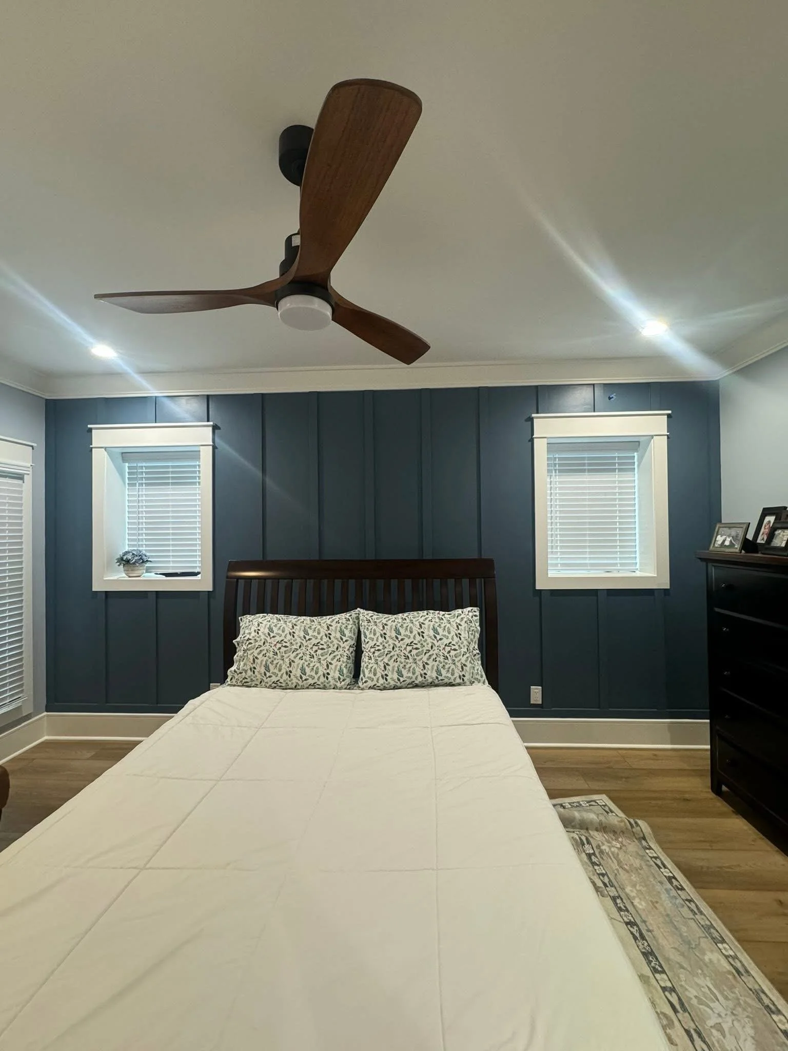 A bedroom with a bed, two windows with blinds, a ceiling fan, and a dresser with pictures.