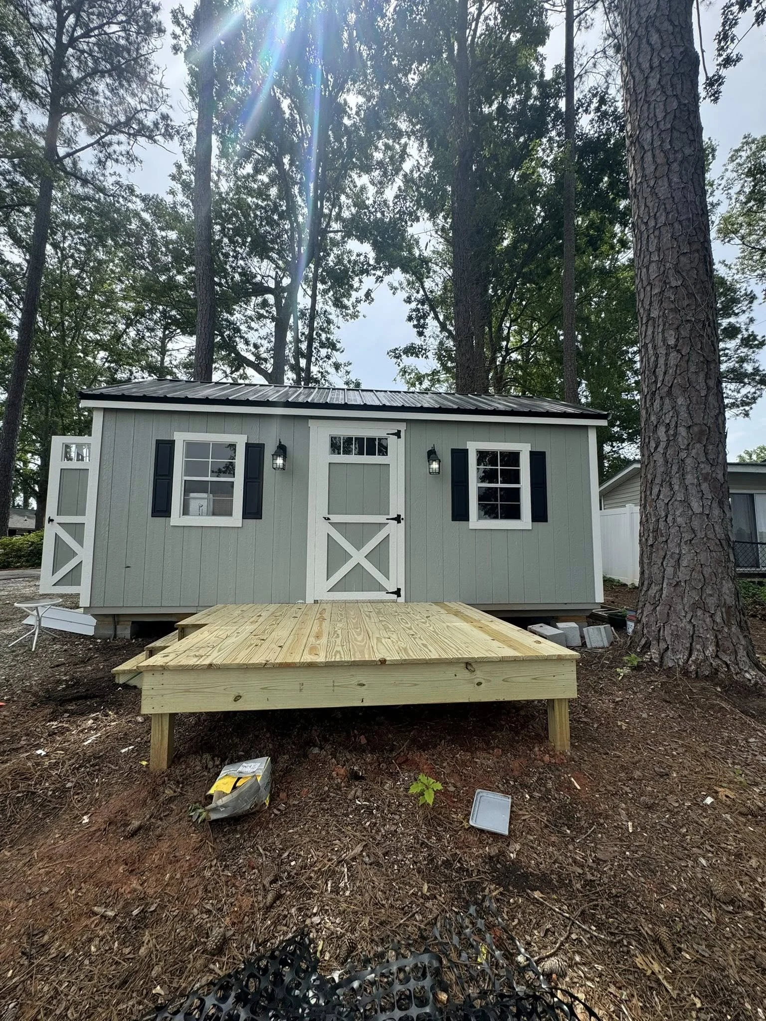 Backyard shed with a new wooden deck, surrounded by tall trees and the sun shining through the branches.