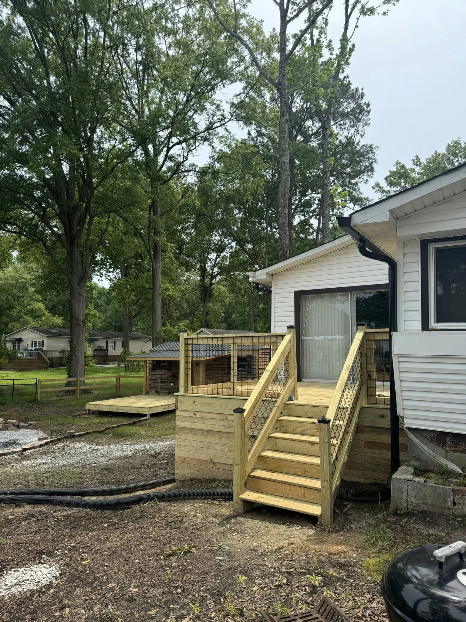 New wooden deck with stairs attached to house, backyard with trees, neighboring houses, and construction materials on the ground.