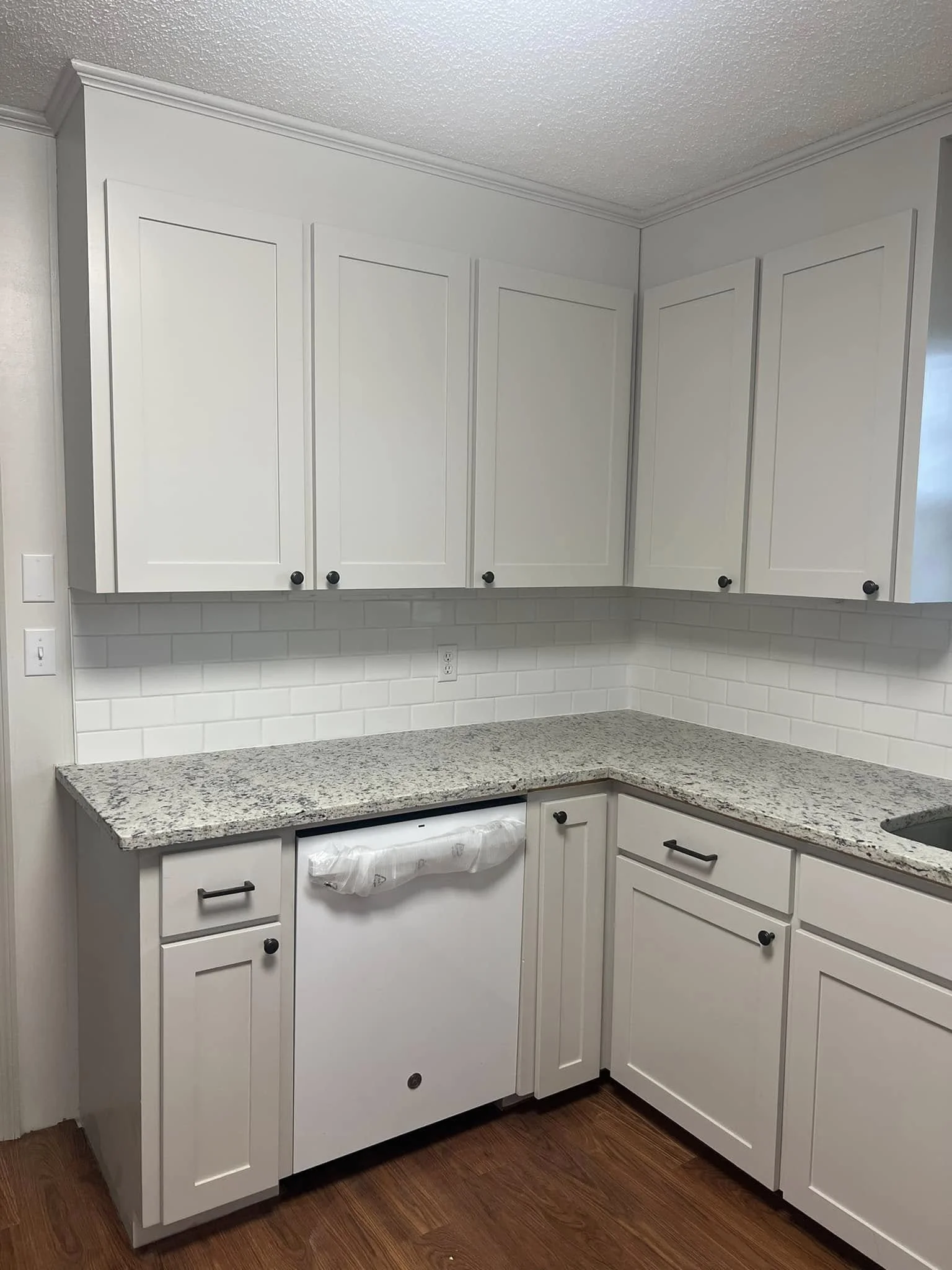 White kitchen cabinets with black knobs, granite countertops, white subway tile backsplash, dishwasher wrapped in plastic, and hardwood floor.