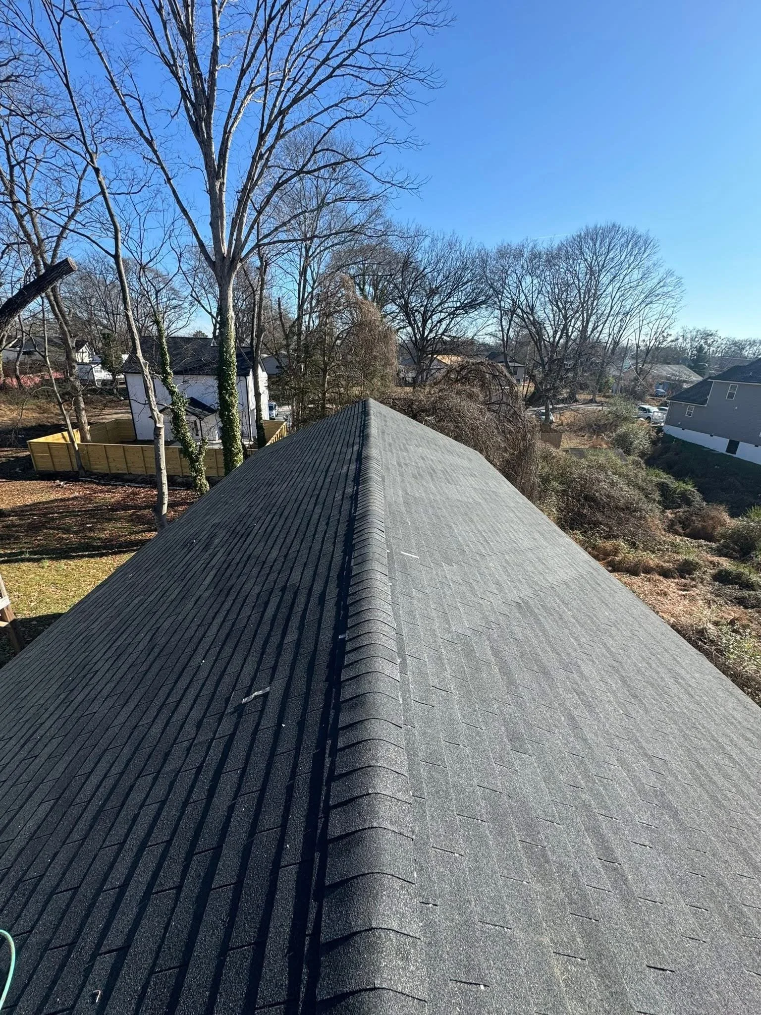 View from the peak of a gray shingled roof with a neighborhood of houses, trees, and a clear blue sky in the background.