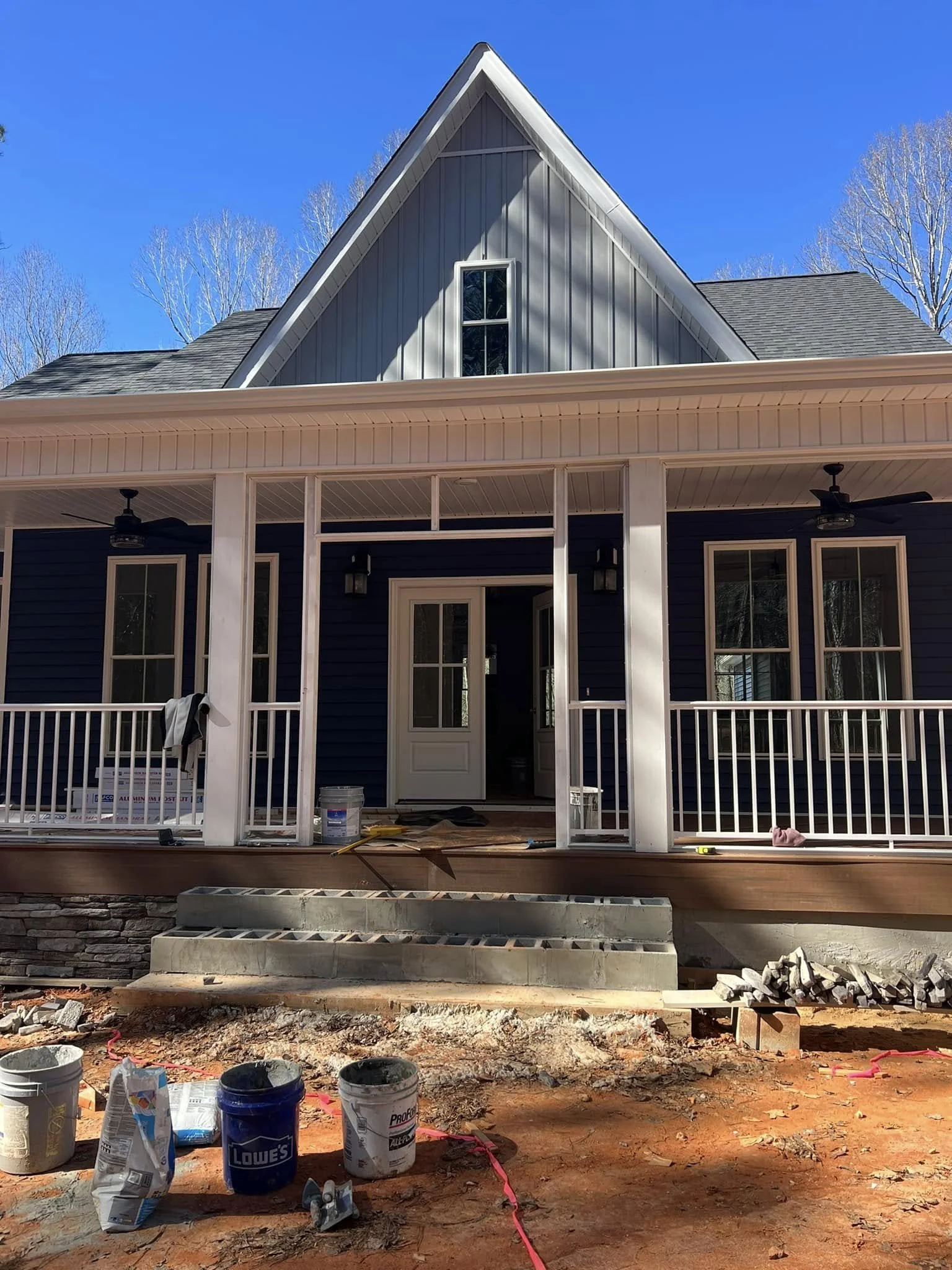 Construction site of a house with a front porch, stairs, and building materials on the ground, in front of a blue house with white trim, under a clear blue sky.