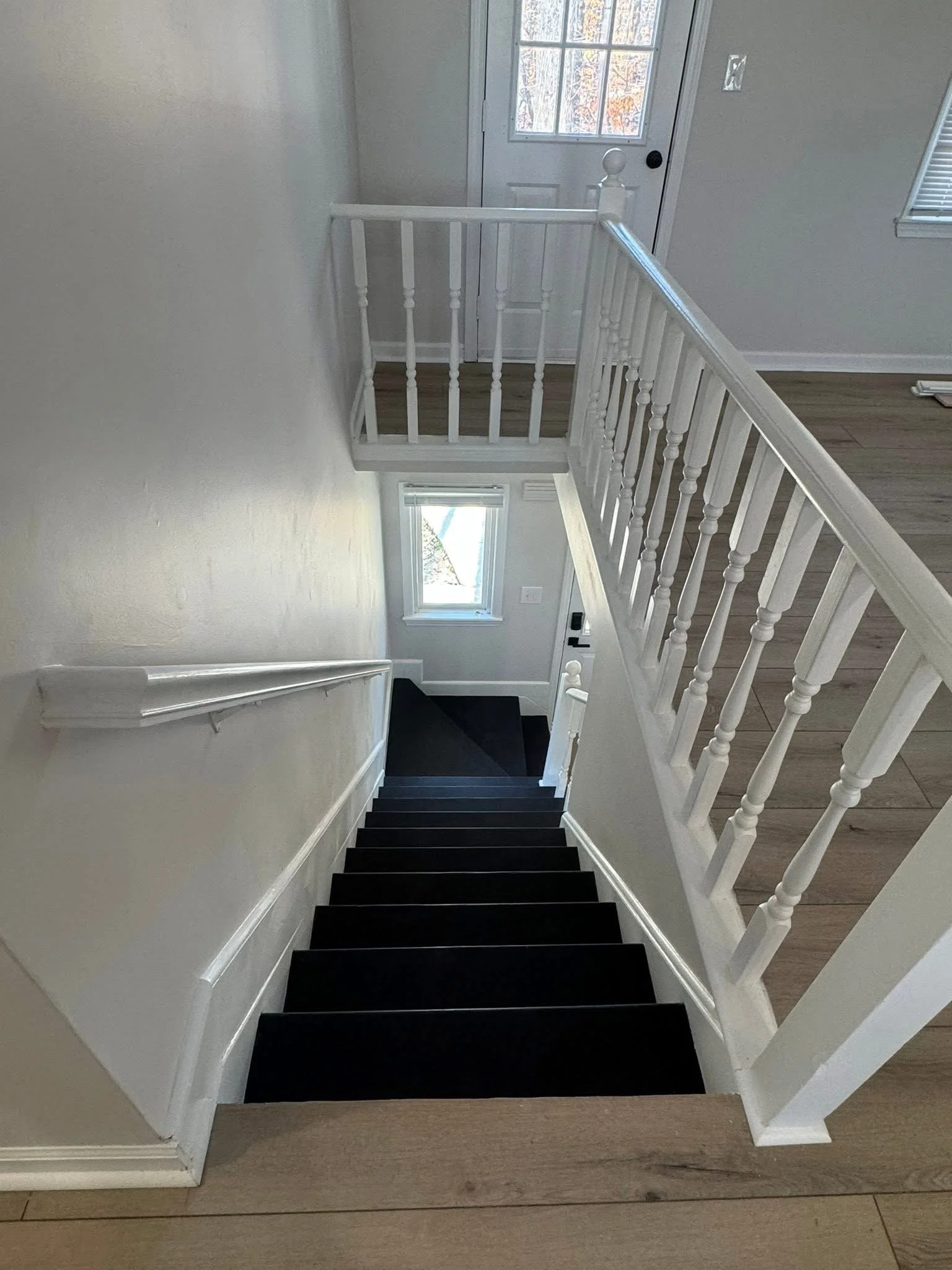 Interior view of a staircase leading down to a doorway with two windows, wooden flooring, white walls, and a white railing.