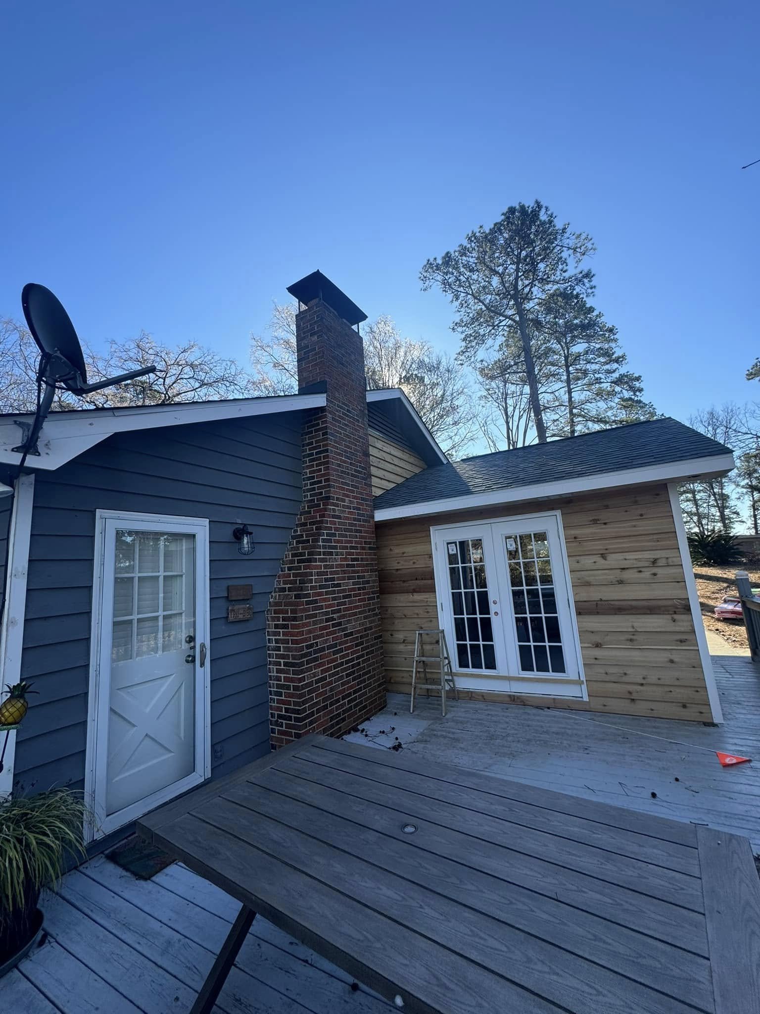 Outdoor patio with wooden deck, blue house with a brick chimney, and sliding glass door. A satellite dish is mounted on the roof, and leafless trees are in the background against a clear blue sky.
