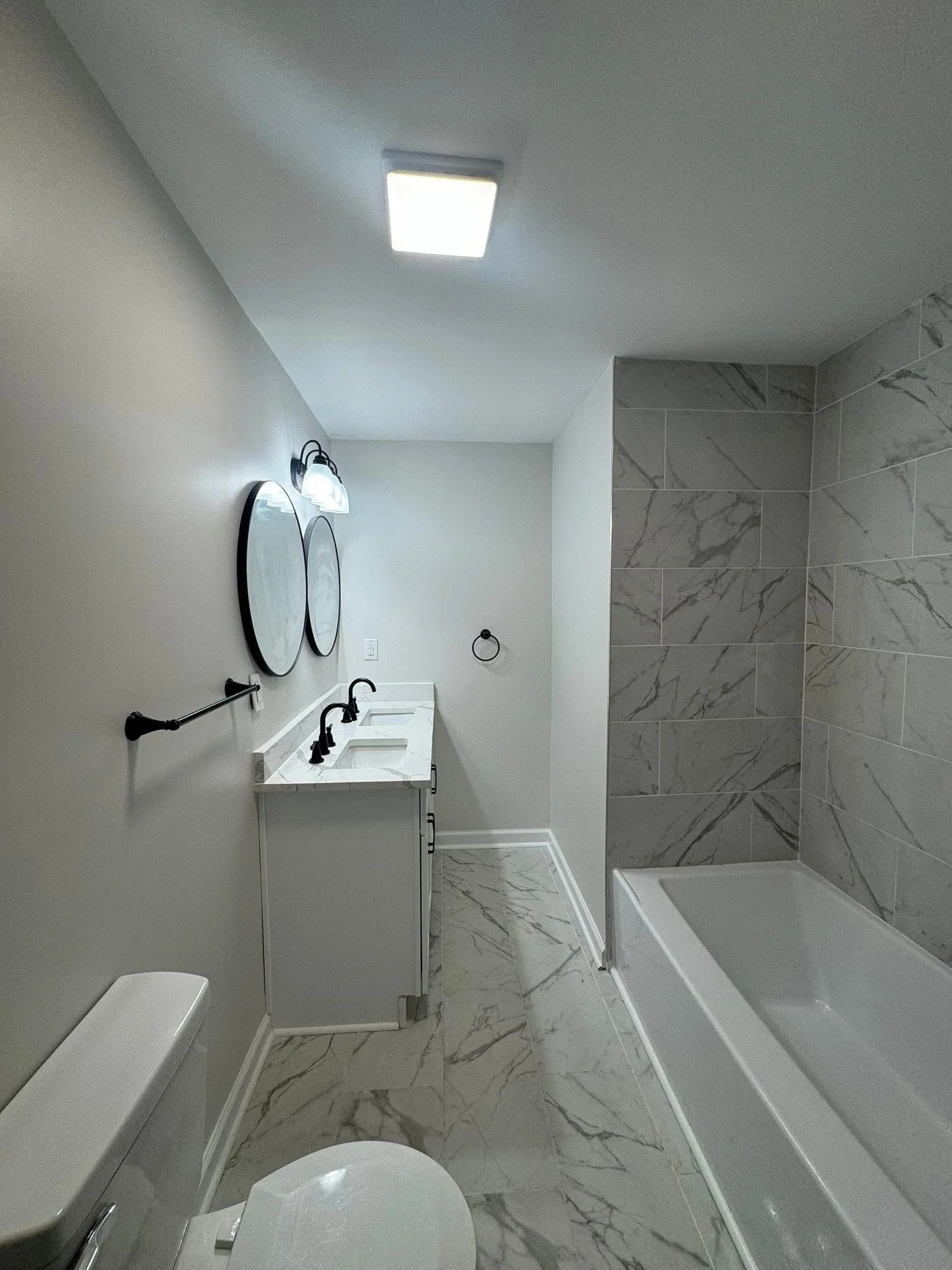 Modern bathroom with white marble floor and shower wall, white vanity with black faucets, two oval mirrors, and black fixtures.