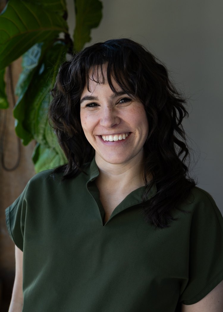 A woman with dark, curly hair smiling in front of a green plant and a neutral background.