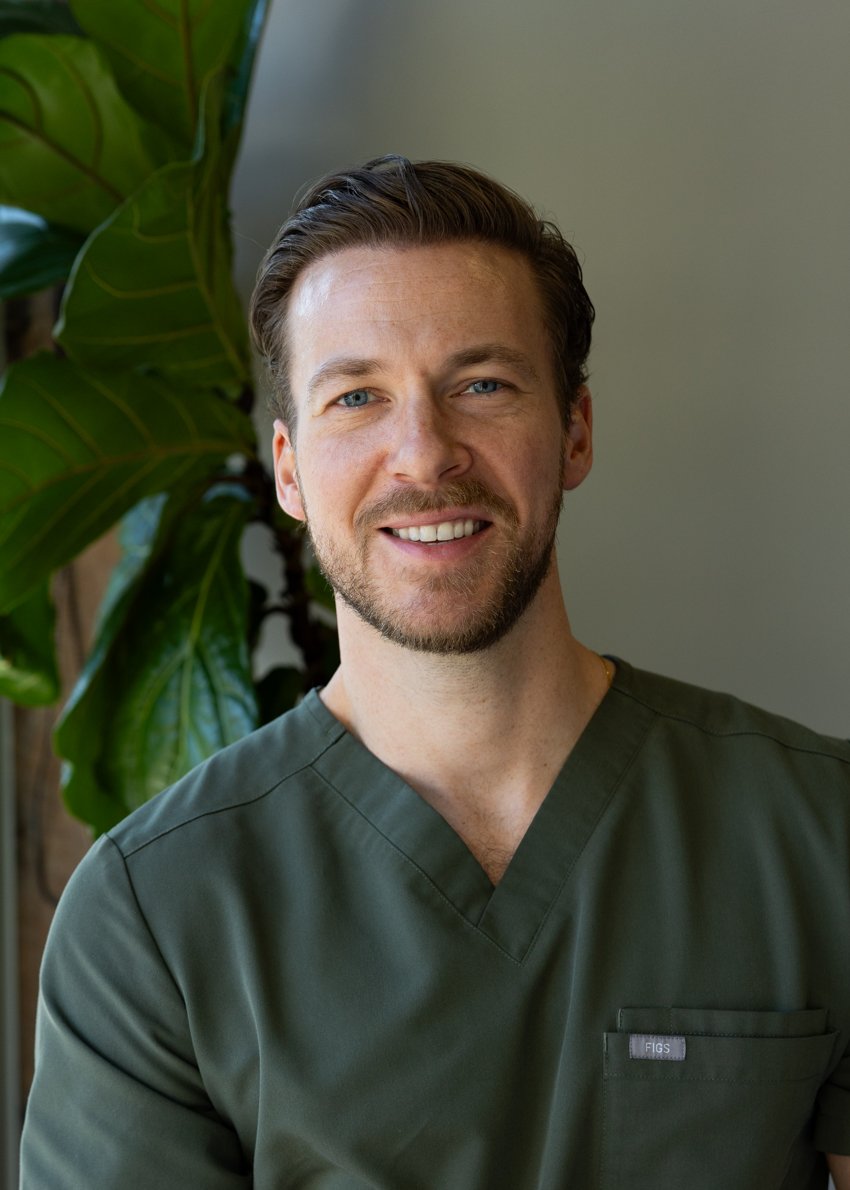 A smiling man with short blonde hair, blue eyes, and a beard, wearing green medical scrubs, standing indoors near a large green leafy plant.