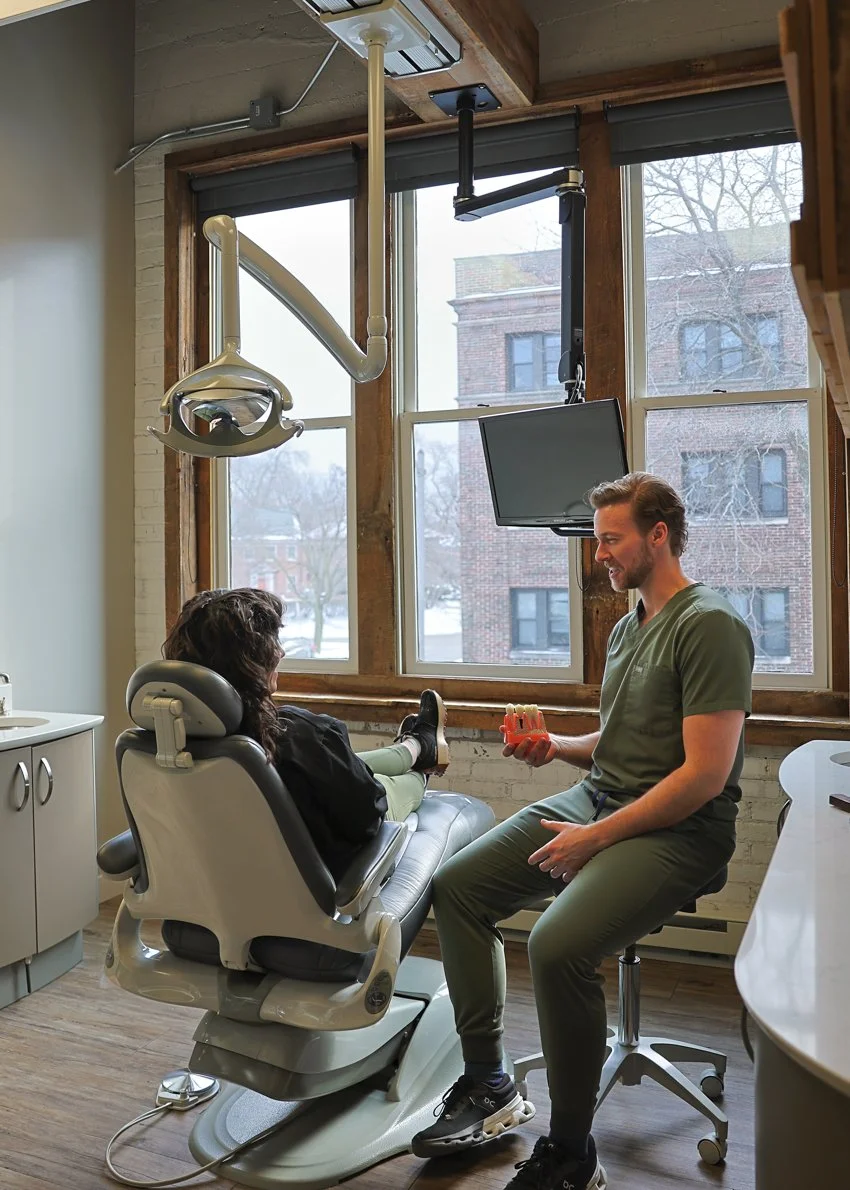 A dentist or dental hygienist speaking with a female patient in a dental office, with dental equipment and a view of buildings and trees outside the window.