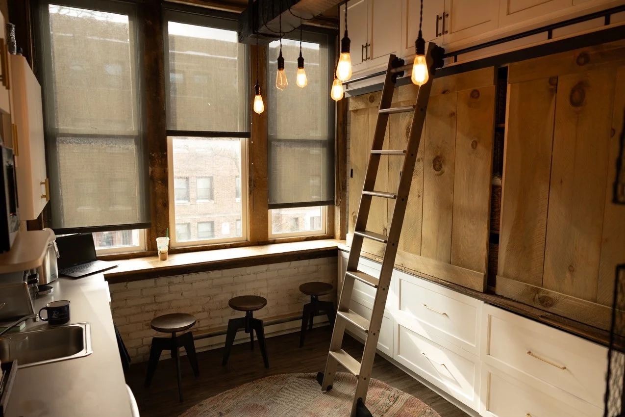 Dental lab space with a ladder, hanging Edison bulbs, white cabinets, a brick wall, and large windows with black blinds.