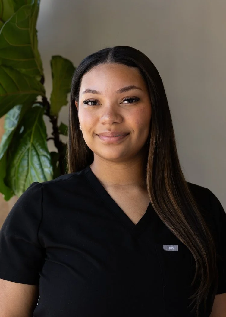 Portrait of a young woman with long dark hair, smiling, wearing a black shirt, standing indoors next to a green plant.