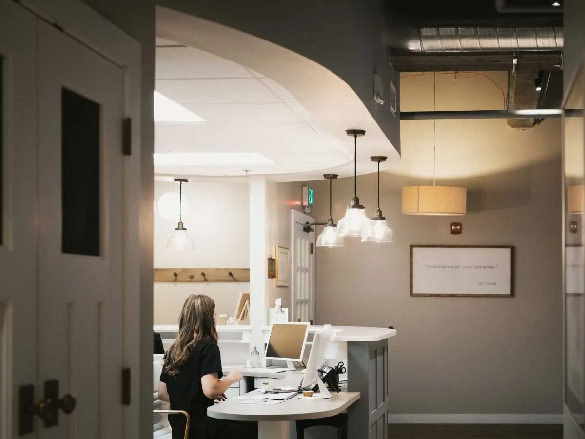 A woman working on a computer at a white reception desk in a modern dental office or, with hanging pendant lights and framed quote on the wall.