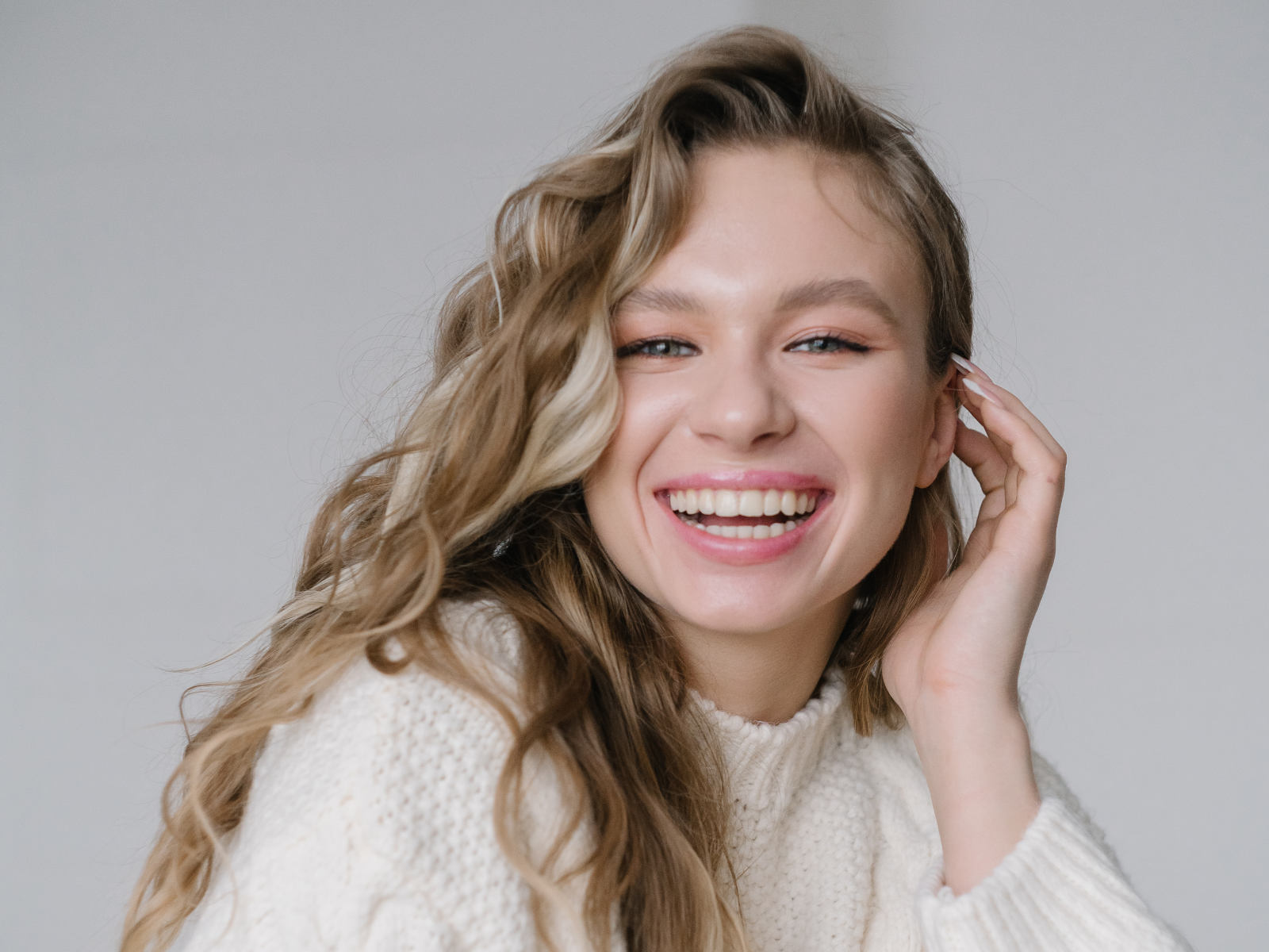 A young woman with wavy, light brown hair smiling and touching her face, wearing a white knit sweater against a plain light background.