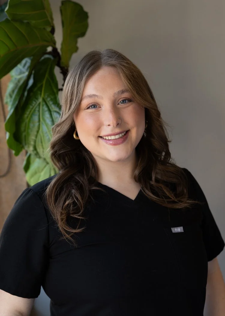 A smiling young woman with wavy brown hair, wearing a black top, gold hoop earrings, standing next to a large green leafy plant.