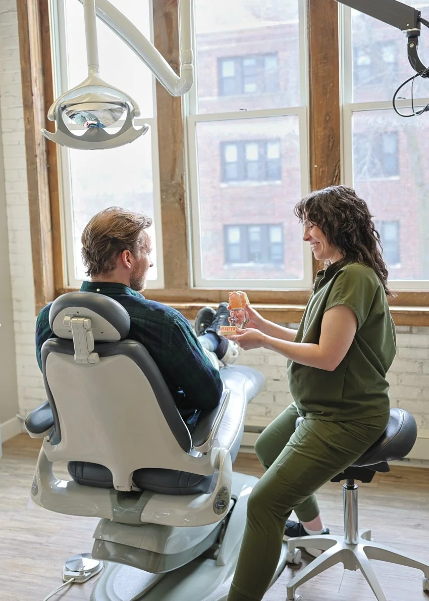 Dentist showing dental model to male patient in dental office with large windows and wooden beams.