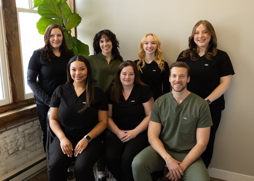 Group of eight healthcare workers, mostly women, posed inside near a window and a large green plant, some in black scrubs, one in olive green scrubs, smiling at the camera.