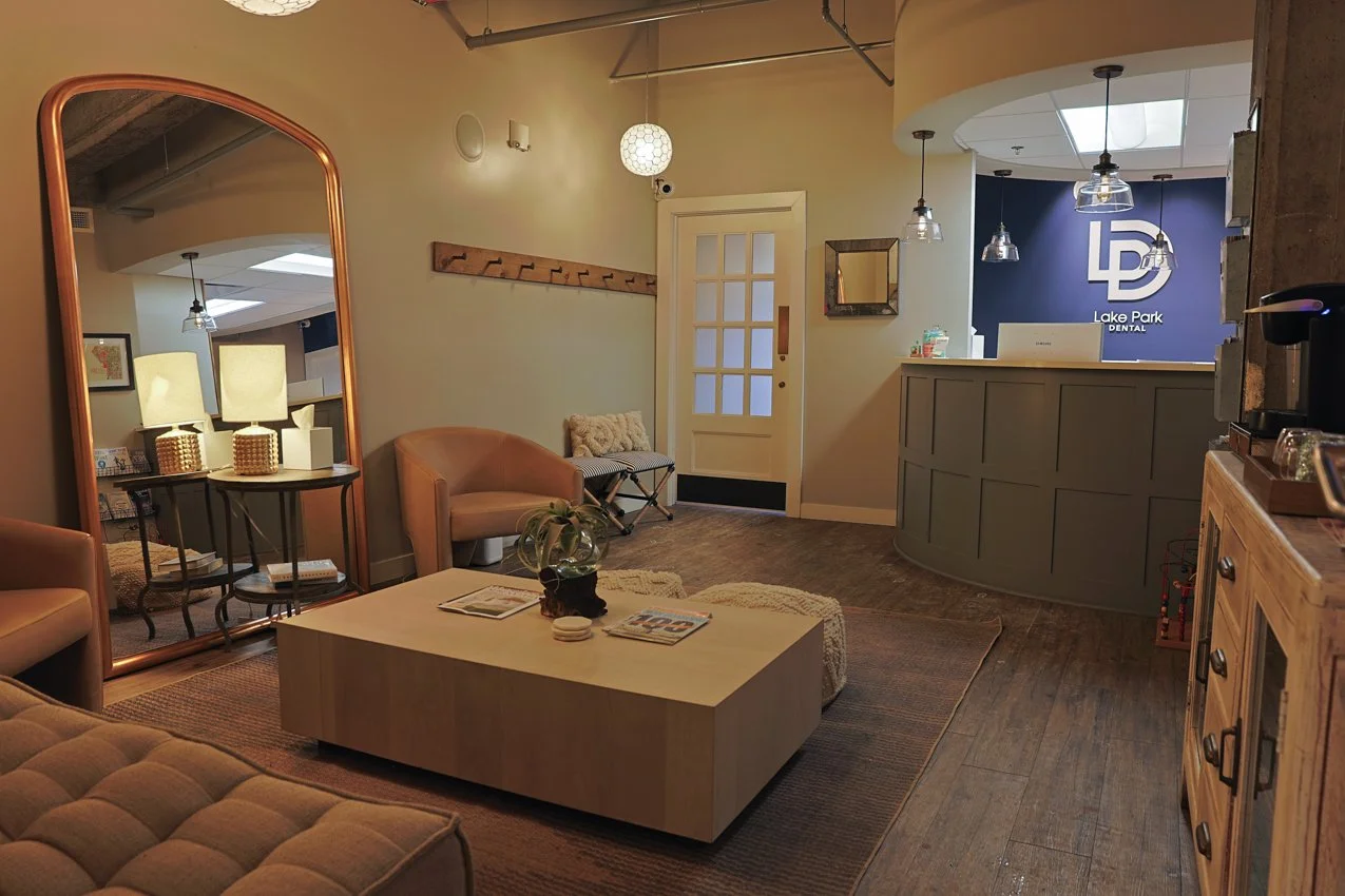 Waiting area of a dental office with beige chairs, a wooden coffee table, mirror, and reception desk with a "Lake Park Dental" sign on a blue wall.