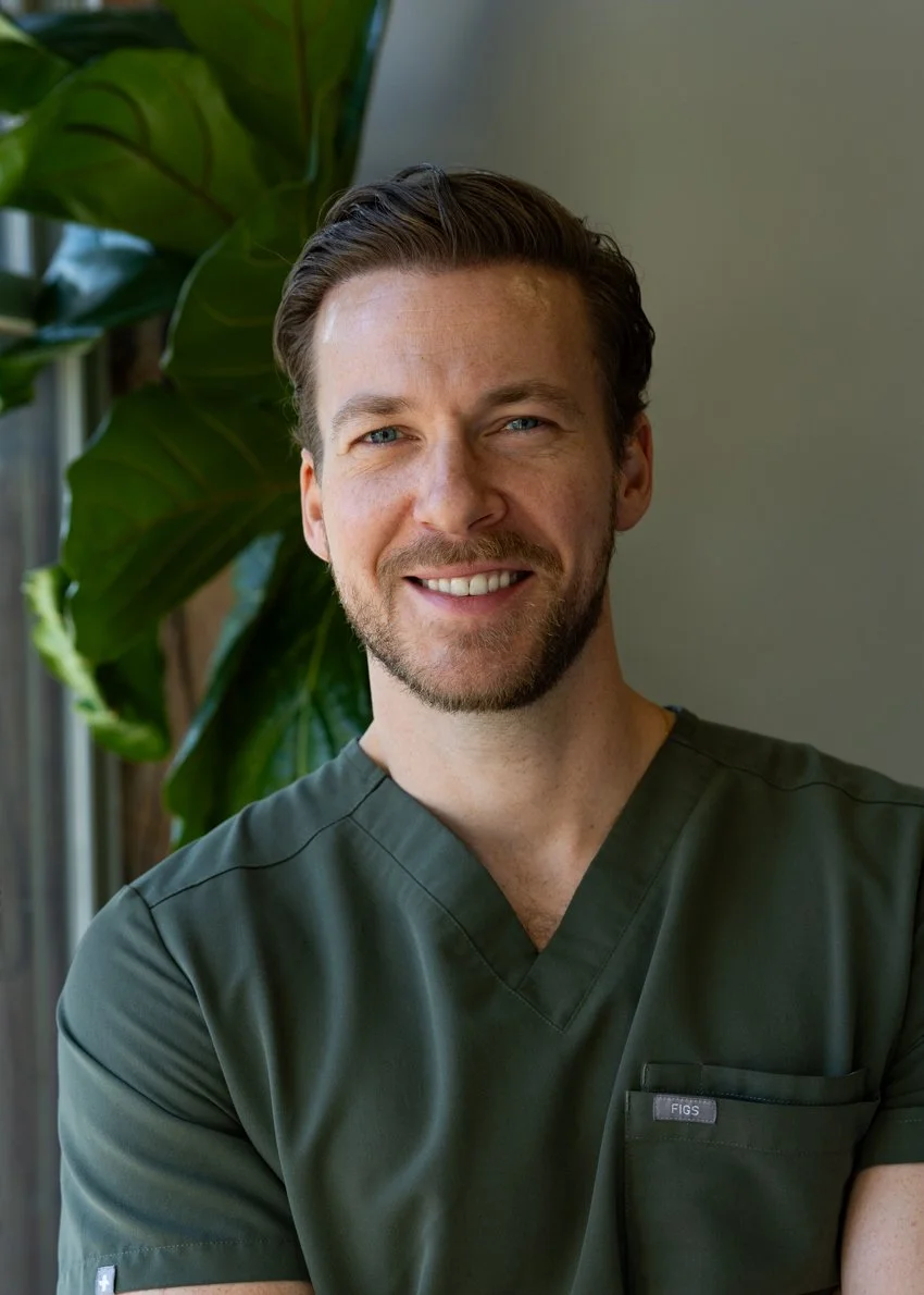 A smiling man with short blonde hair and a beard, wearing green medical scrubs, sitting near a window with large green leaves in the background.