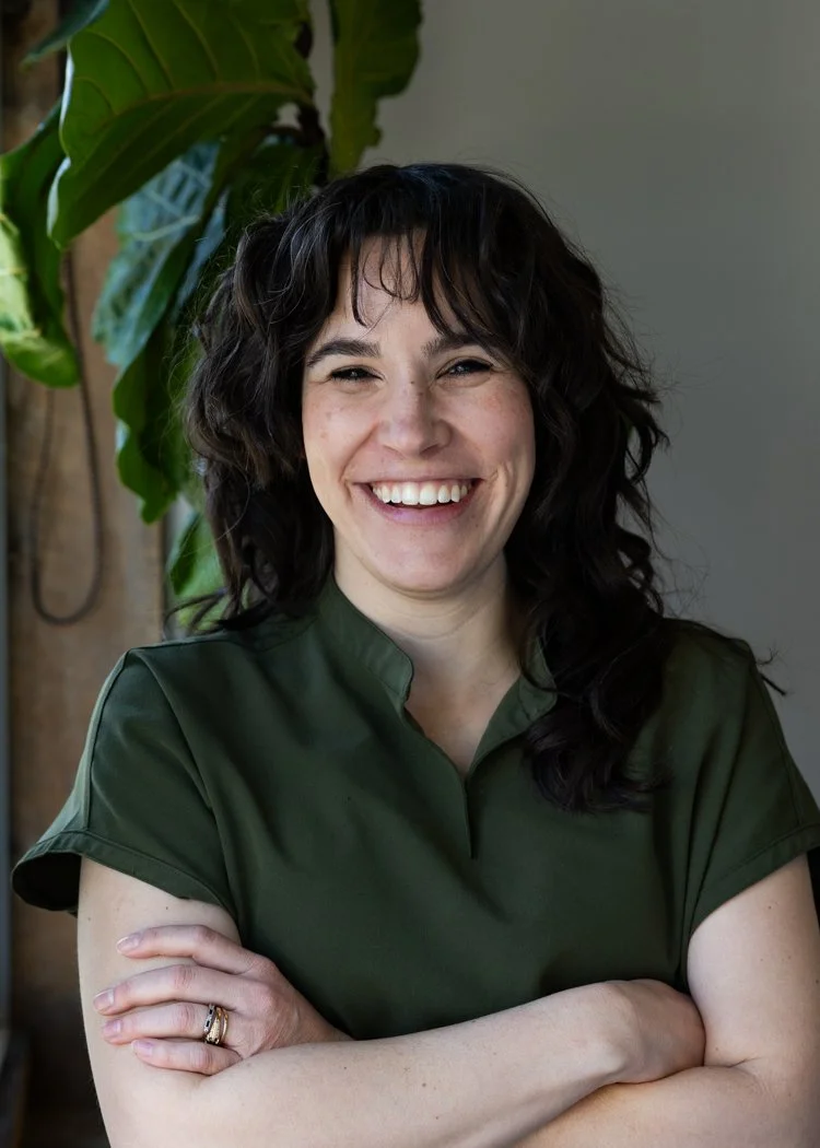 A woman with dark, wavy hair smiling with arms crossed, wearing a dark green top, standing in front of a leafy green plant.
