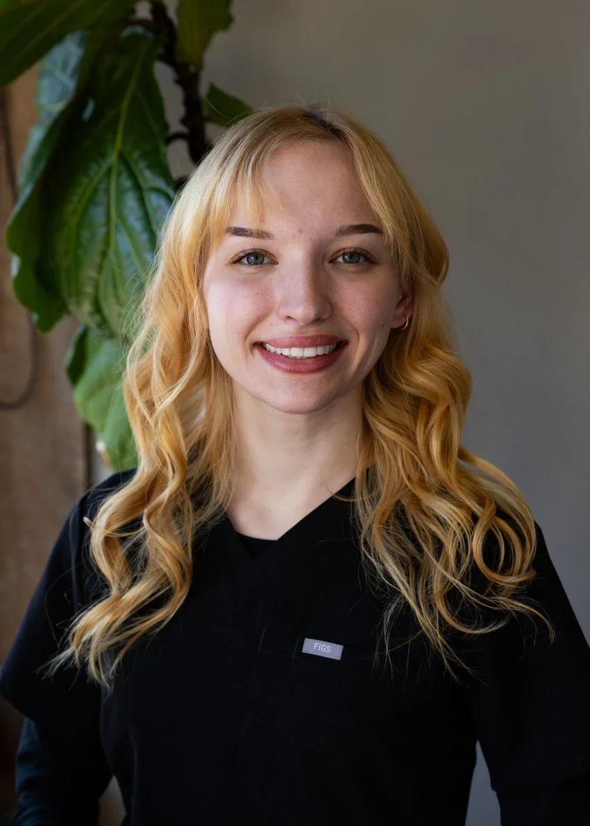 A young woman with blonde, wavy hair and a black shirt standing indoors near a green leafy plant.
