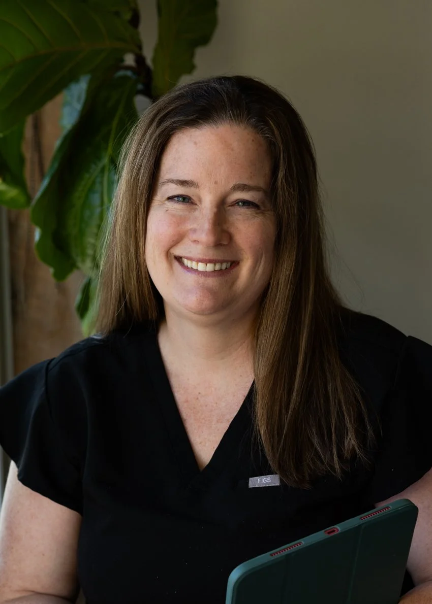 A woman with long brown hair smiling in front of a large green plant, wearing a black top and holding a tablet.