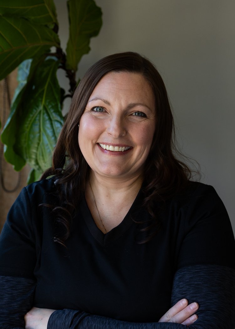 A woman with long dark hair wearing a black top, smiling with arms crossed, standing near a large green plant indoors.