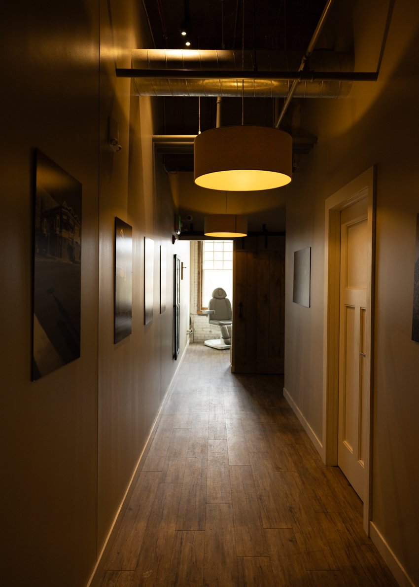 A narrow hallway with wood flooring and beige walls decorated with framed pictures. The hallway is lit by ceiling-mounted circular lights and leads to a bright room with a window at the end, where an adjustable office chair is visible.