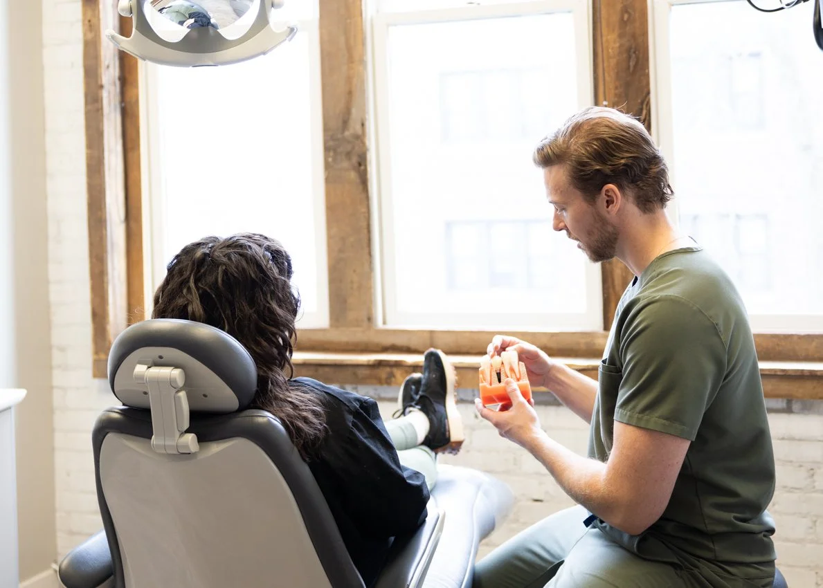 Dentist in green scrubs consulting with a female patient in a dental office with large windows and wooden framing.