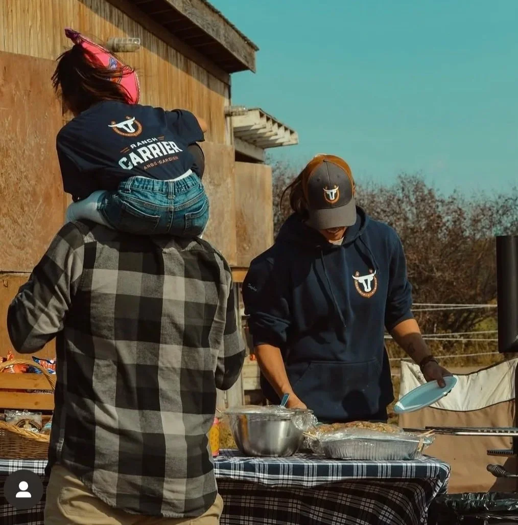 Trois personnes à une table avec de la nourriture en extérieur, deux portent des casquettes et un porte un casque, un d'eux porte un sweat à capuche et un autre un t-shirt avec un logo.
