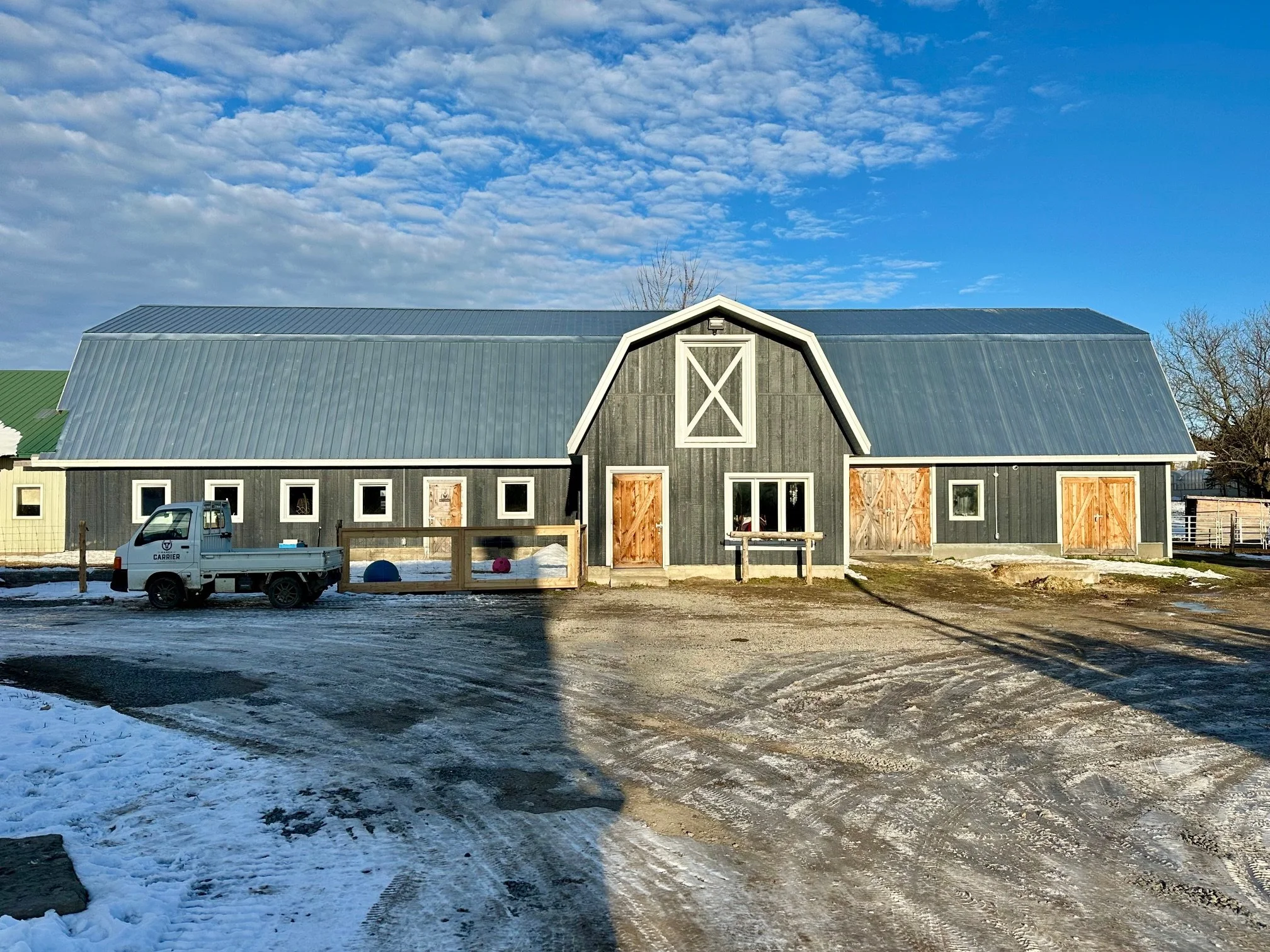 Une grande grange en bois peint en gris, avec un toit métallique, située dans un paysage enneigé lors d'une journée ensoleillée.