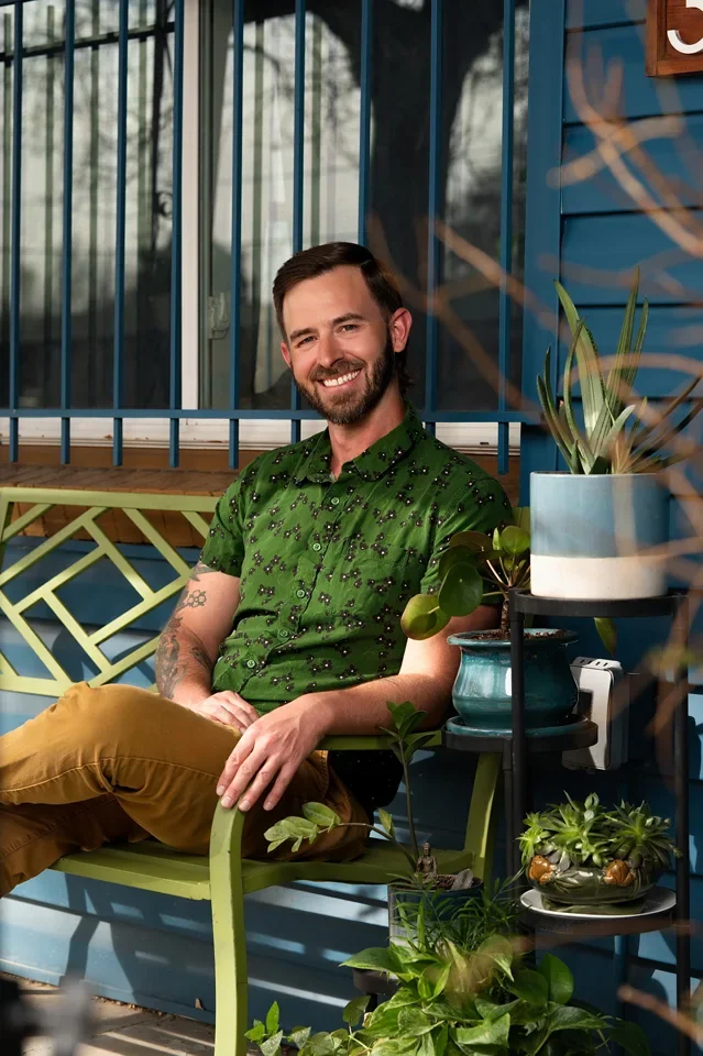 Portrait of Colorado Licensed Professional Counselor, Adam Stanford, with short dark hair wearing a green patterned shirt and mustard-colored pants sitting on a green bench in front of a blue wall with plants around.