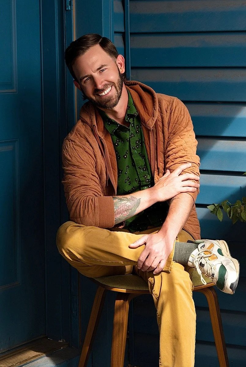 Portrait of Licensed Professional Counselor, Adam Stanford, posing in Colorado. He wears a green shirt, a brown jacket, mustard-colored pants, and white sneakers.