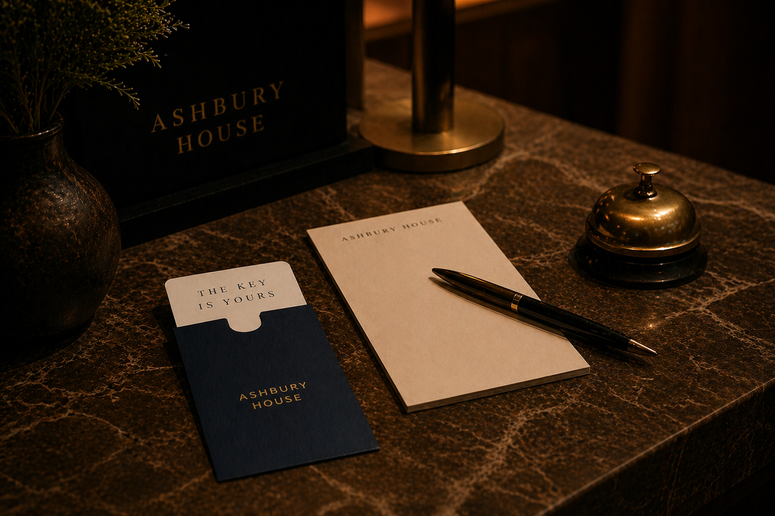 A marble reception desk with a black and gold sign that says 'Ashbury House,' a small vase with greenery, a guest check-in envelope, a notepad, and a pen.