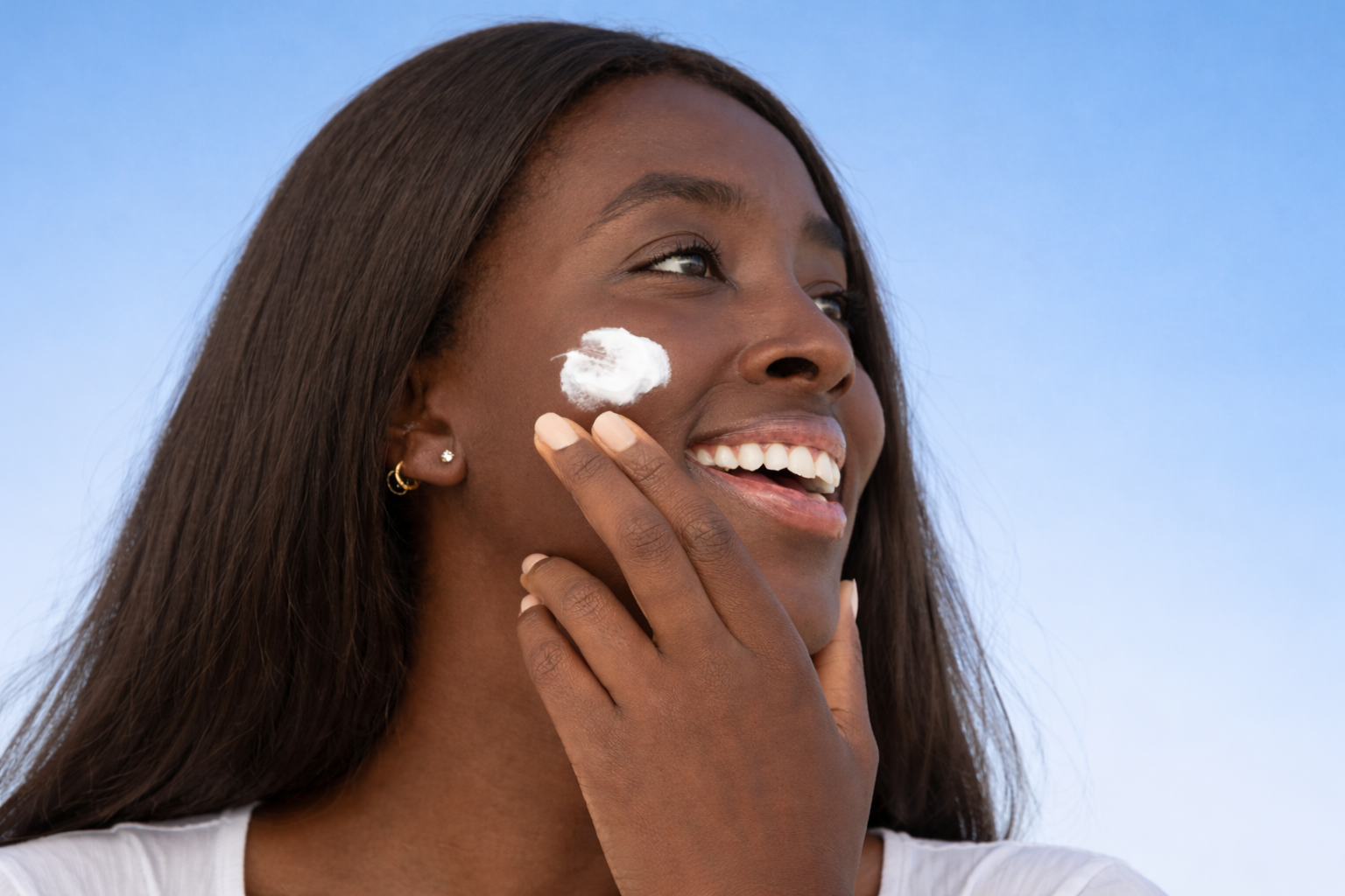 A woman smiling outdoors with the sky in the background, applying sunscreen to her face.