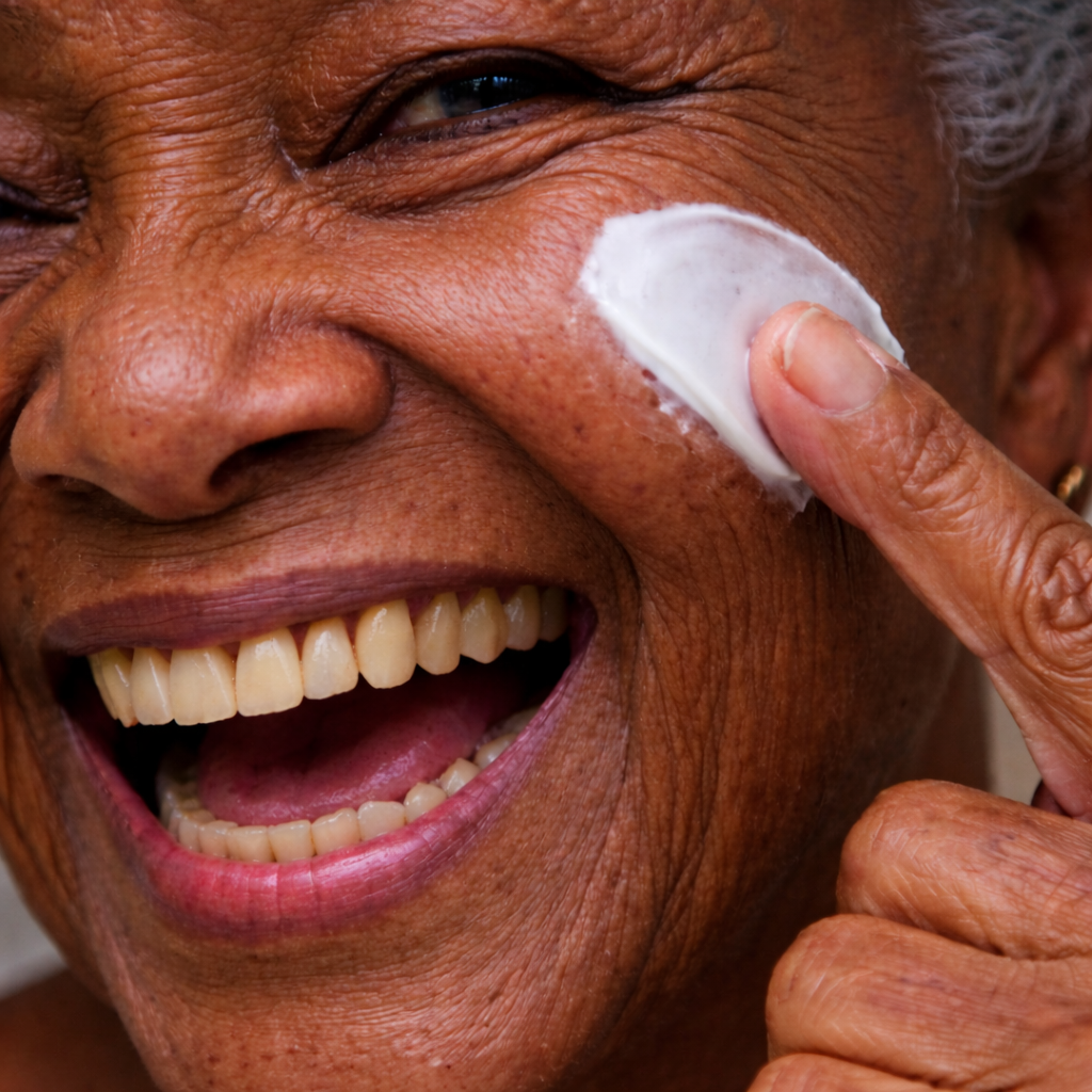 Close-up of an elderly woman with a big smile, applying white cream or lotion to her nose with her finger.