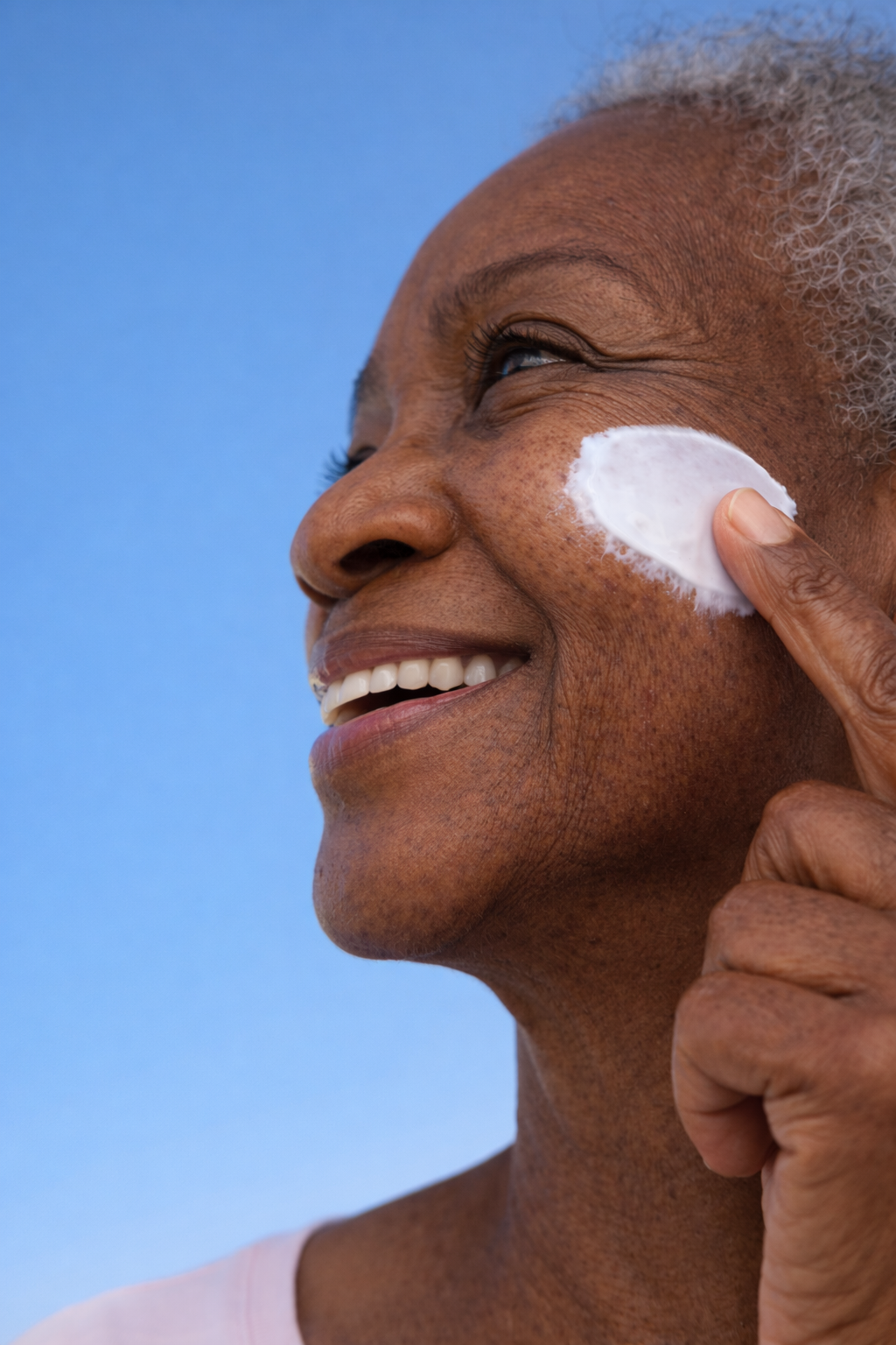 Close-up of smiling elderly African American woman applying sunscreen to her face against a blue sky background.