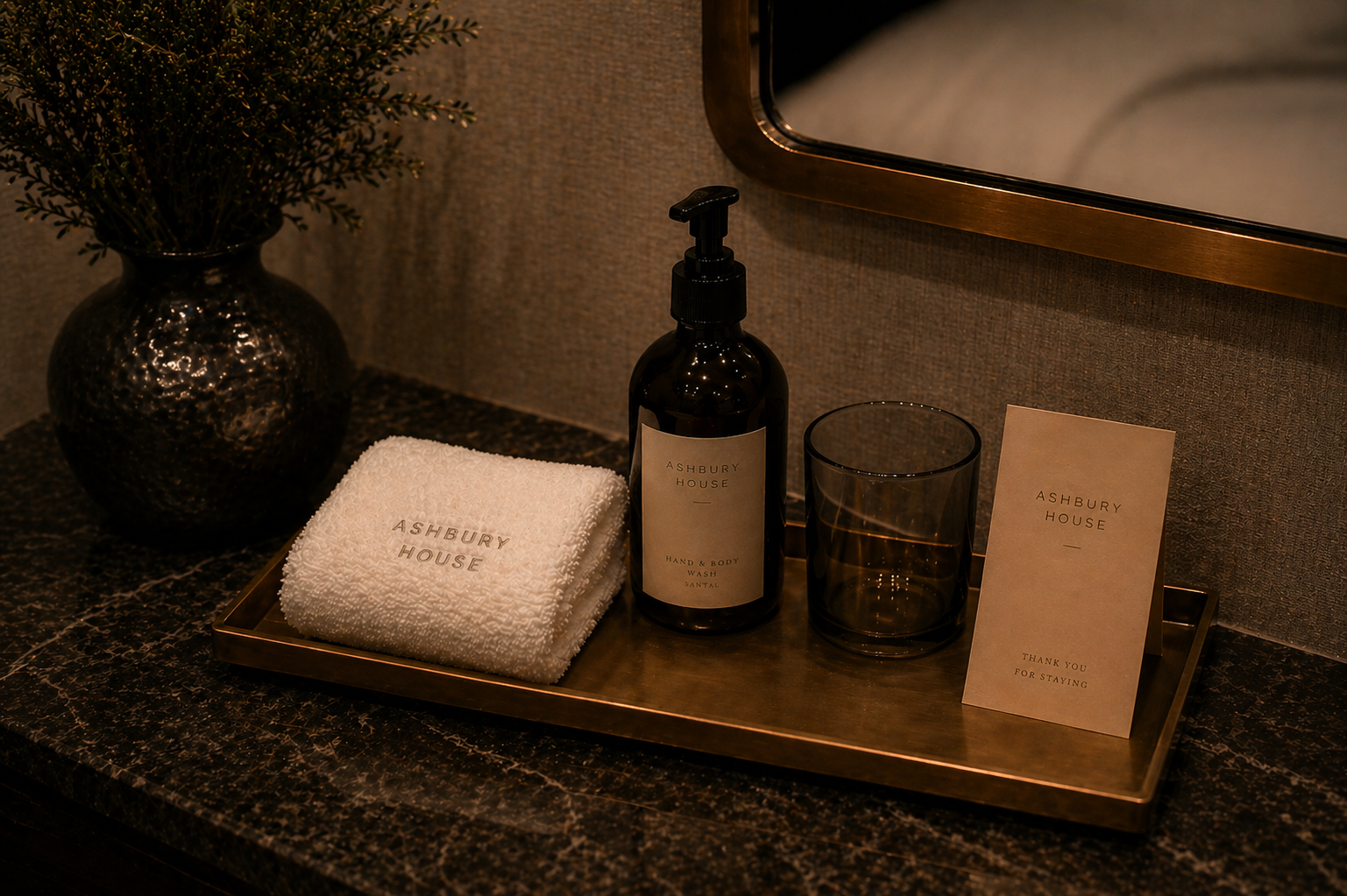A bathroom tray with a black soap dispenser, a glass, a folded white towel embroidered with 'Ashbury House', and a thank you note. A black decorative vase with greenery is in the background.