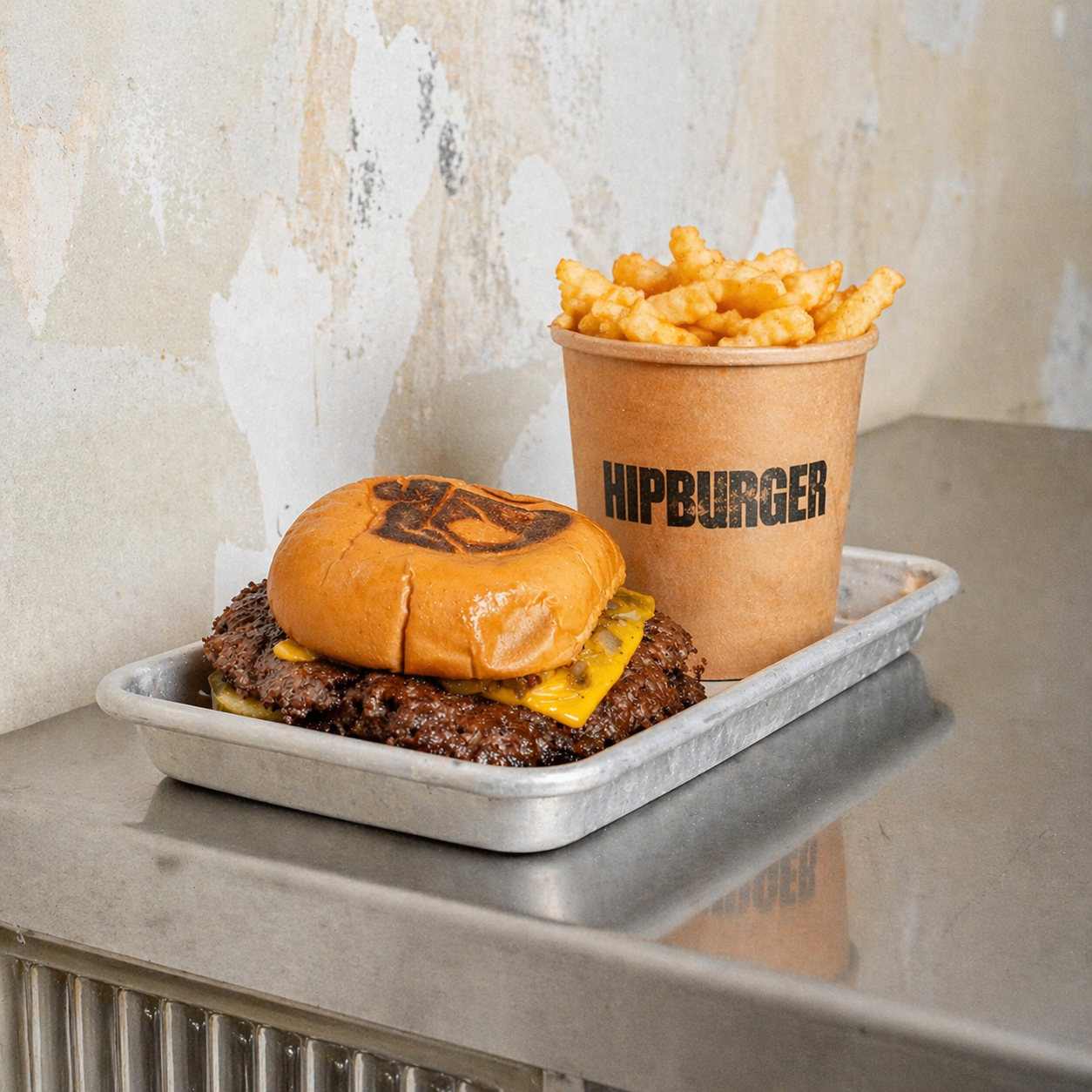 A tray with a cheeseburger topped with pickles, cheddar cheese, and a bun, alongside a paper cup filled with French fries, all on a metal surface against a textured wall.