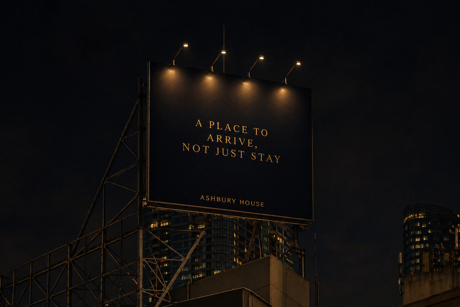 Night-time city billboard with a quote that reads 'A place to arrive, not just stay' and 'Ashbury House' at the bottom, lit by four spotlights.
