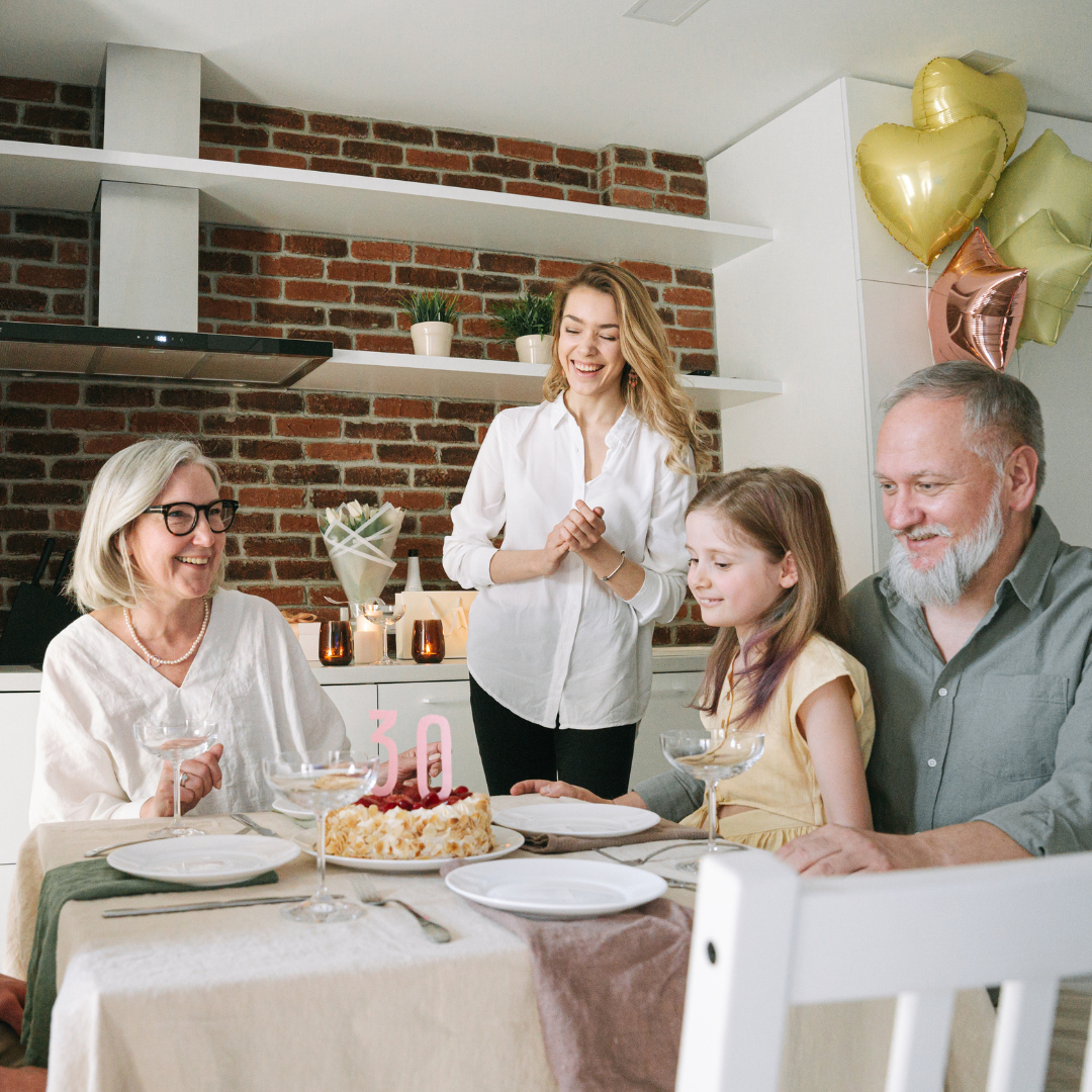 Una reunión familiar en celebración de cumpleaños con una mujer, un hombre, una niña y una mujer mayor sentados en una mesa, decorada con un pastel y globos dorados y rosados, en un ambiente acogedor de cocina con paredes de ladrillos y estanterías blancas.