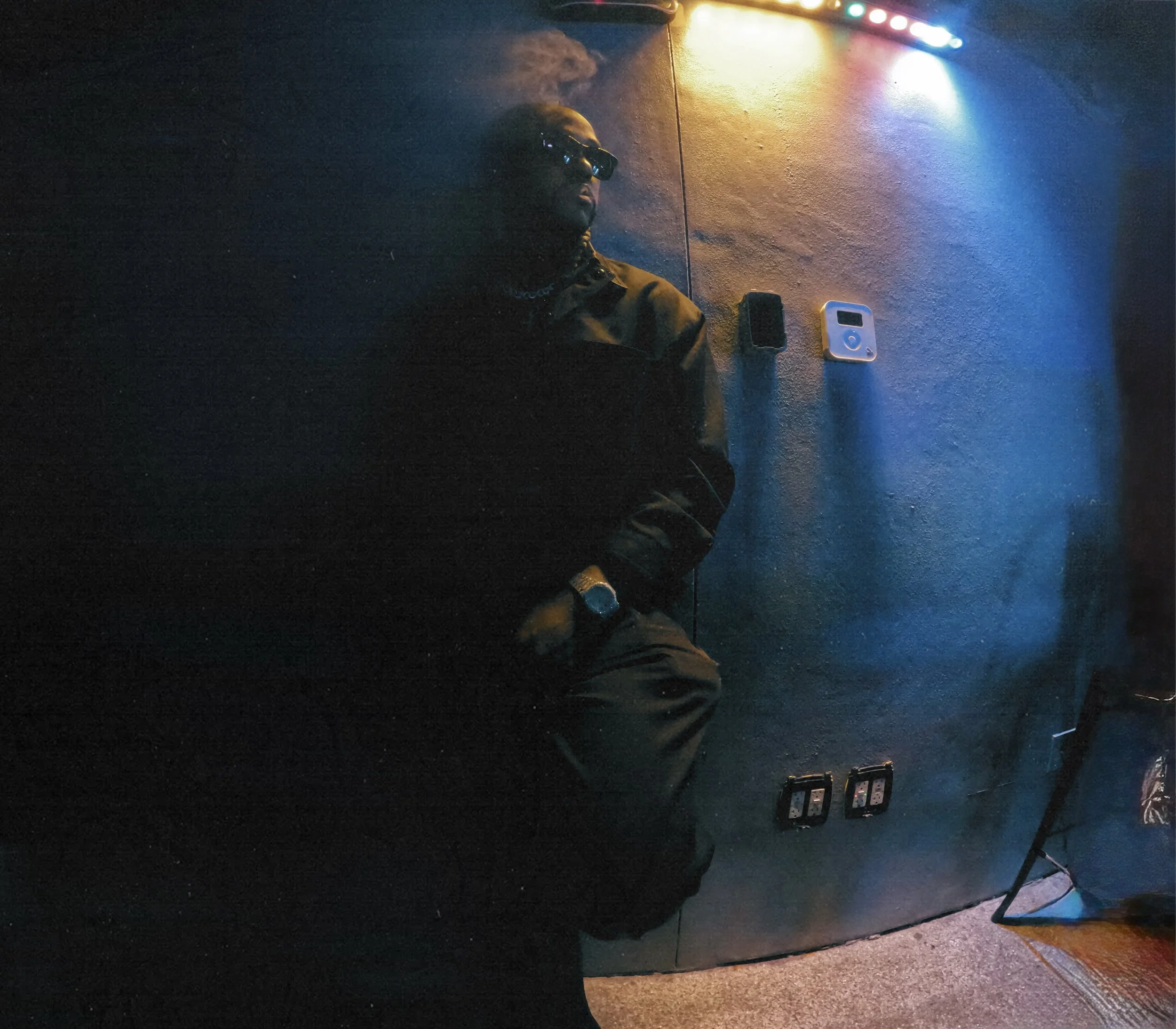 A person leaning against a dark wall, wearing sunglasses, a black jacket, and a watch, with smoke coming from their mouth and colorful lights above.