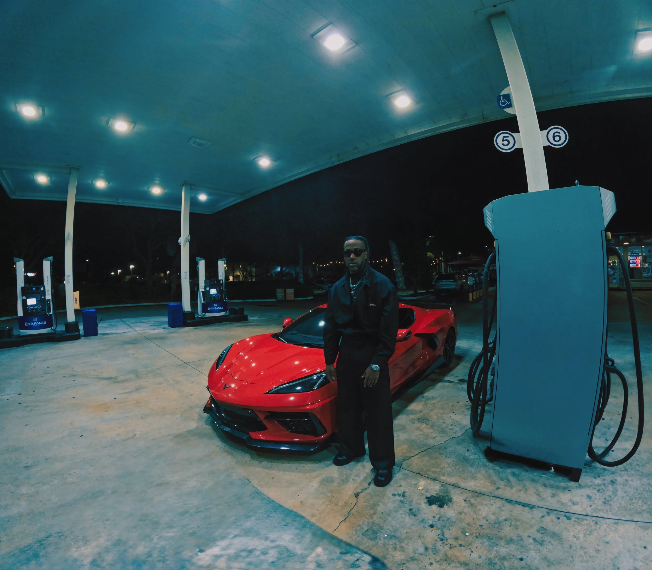 A man in black clothing and sunglasses standing next to a red sports car at a gas station at night.