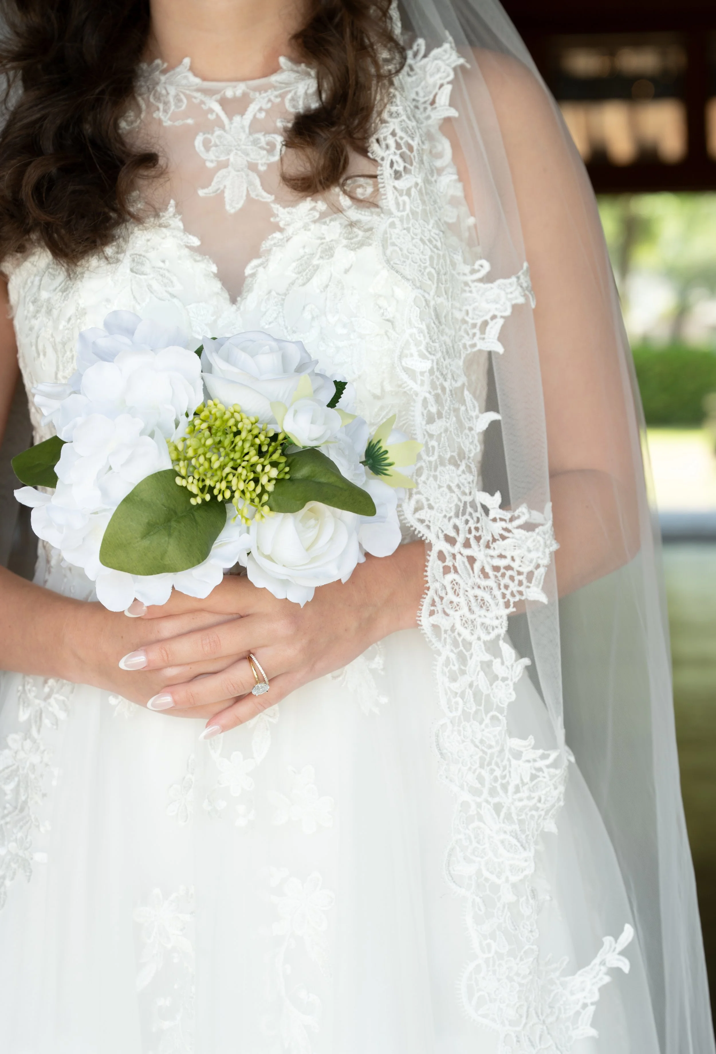 Close-up of a bride holding a bouquet of white flowers, wearing a lace wedding dress, with a wedding ring on her finger.